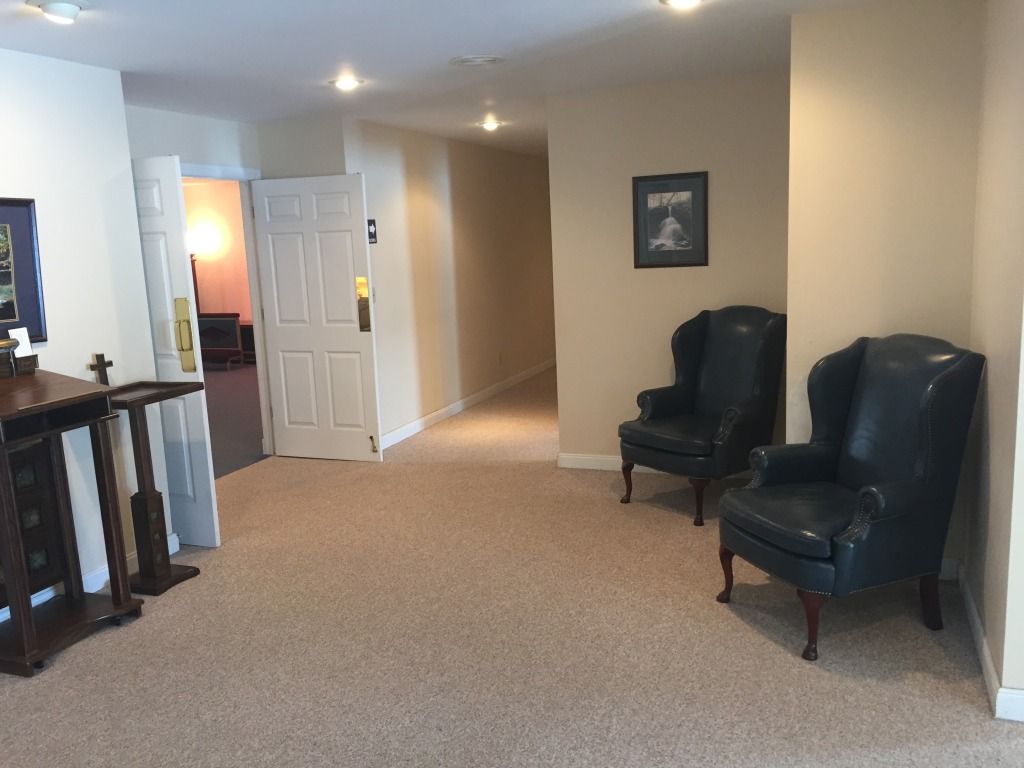 A hallway with tan carpet and walls. Two dark leather armchairs sit near a framed picture. A door is ajar.