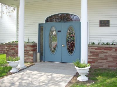 Blue double doors with oval stained glass, flanked by columns and planters, leading into a white building.