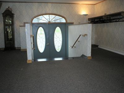 Gray double doors with oval stained glass, leading to a room with a grandfather clock, carpet, and a ramp with a handrail.
