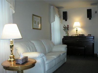 A living room with white sofa, lamps, piano, and speakers on the wall.
