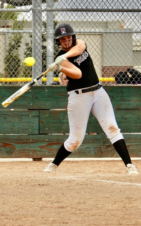 A softball player in a black jersey and white pants swings a bat at a yellow ball on a dirt field behind a fence.
