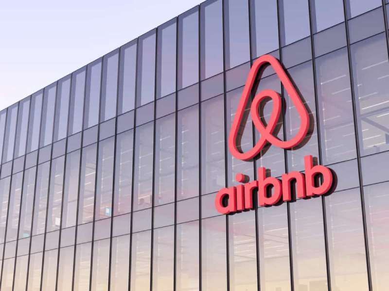El logotipo de Airbnb en la fachada de vidrio de un edificio moderno, rojo sobre blanco, con un cielo azul claro.