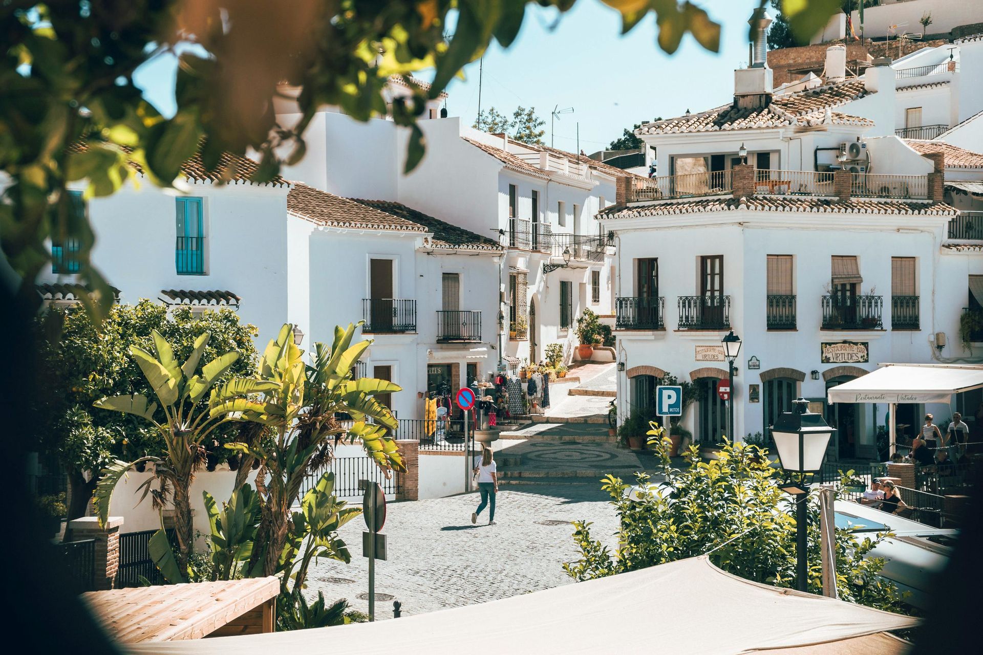 Una plaza soleada con edificios blancos, calles empedradas y exuberante vegetación enmarcada por hojas en primer plano.