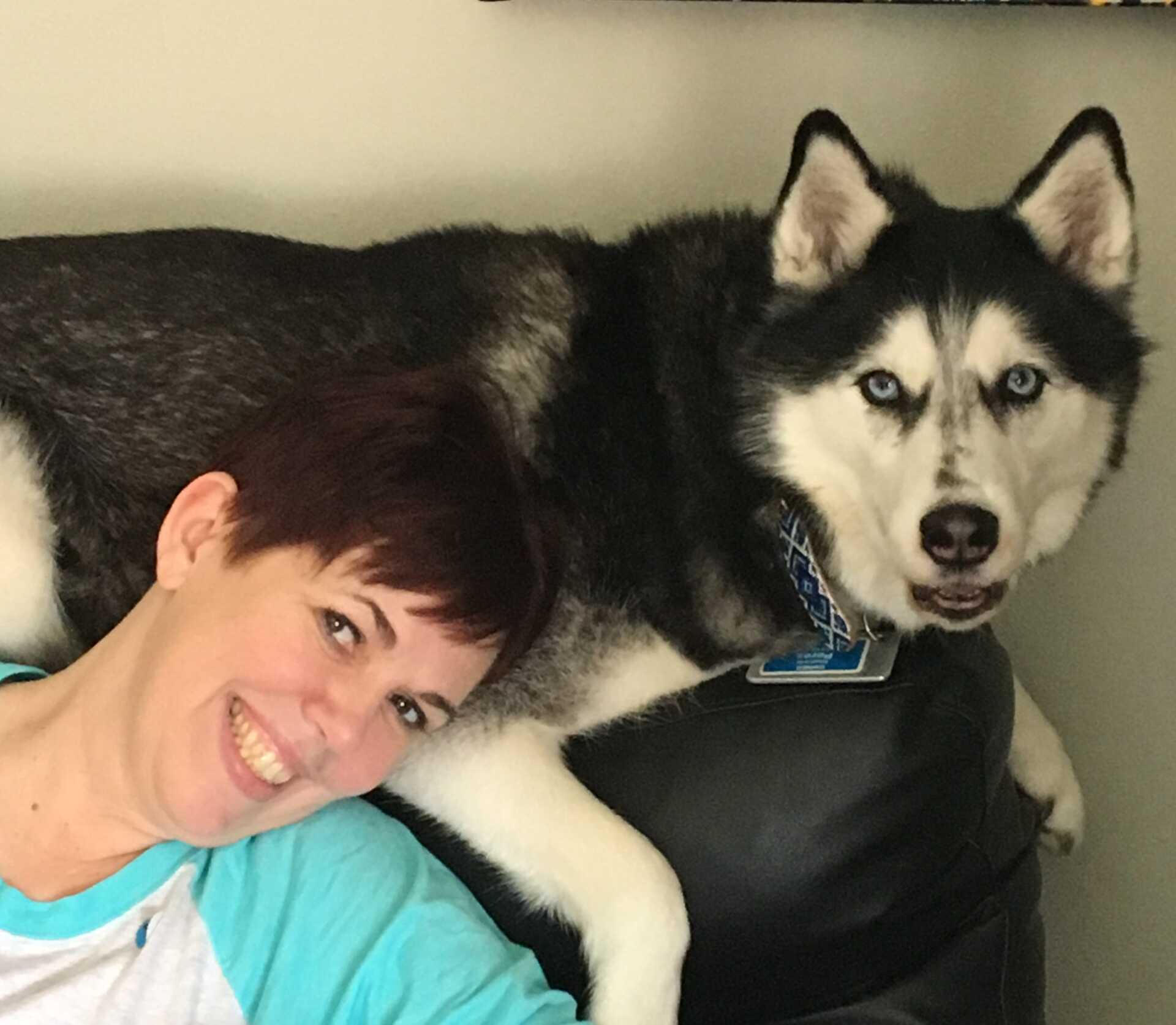 A woman is laying on a couch next to a husky dog