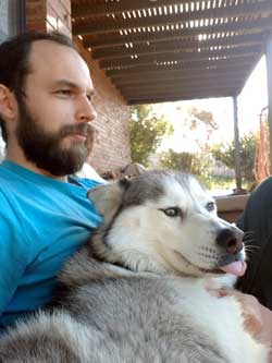 A man with a beard is holding a husky dog on his lap.