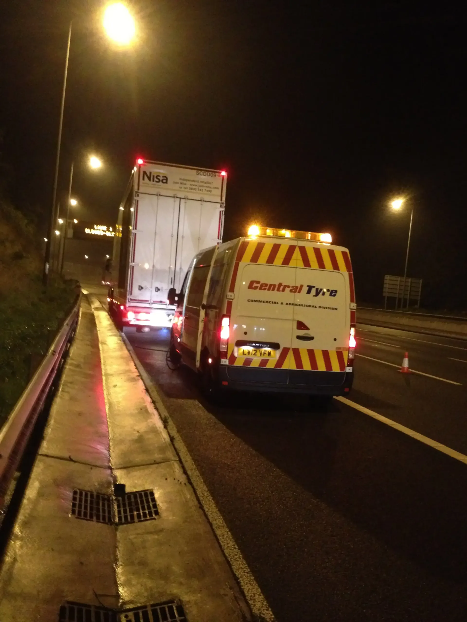 A Central Tyre service van with flashing lights next to a stalled truck on the side of a road at night.
