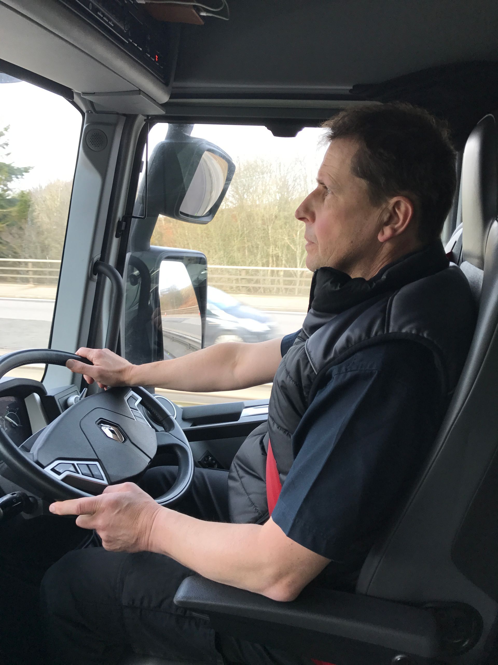 Man driving a truck, hands on the steering wheel, looking forward. Inside the cab, day.