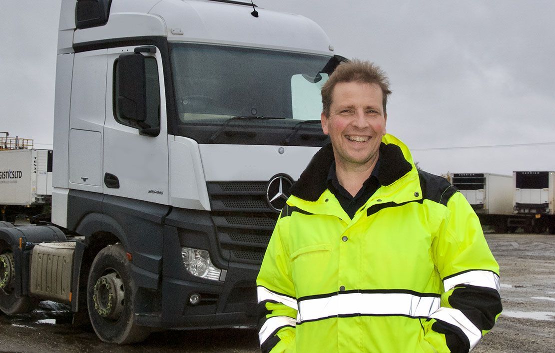Man in yellow jacket smiles next to a white truck. Outside, overcast.