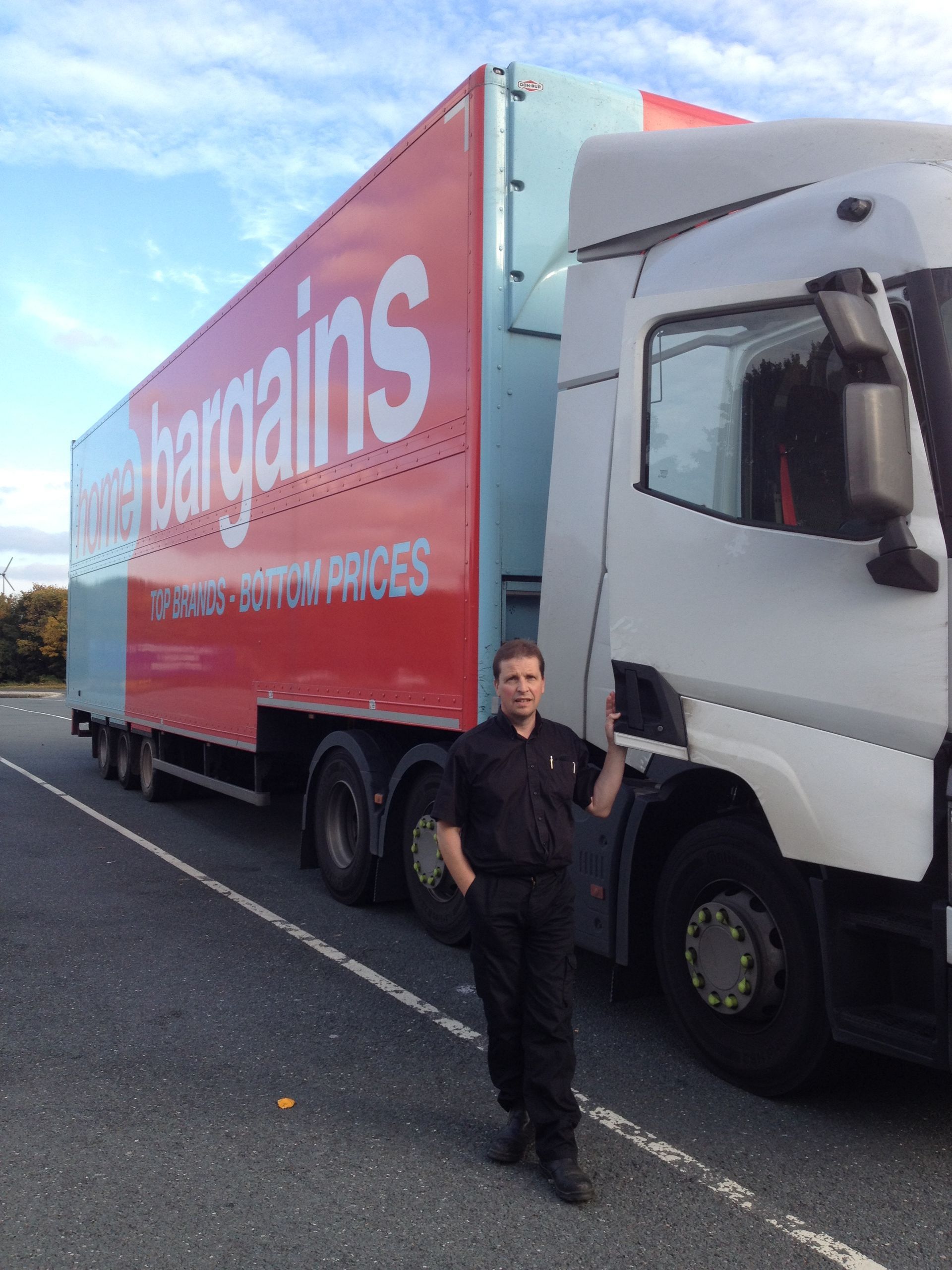 Man standing next to a Home Bargains truck. The truck is white and red. Blue sky in the background.