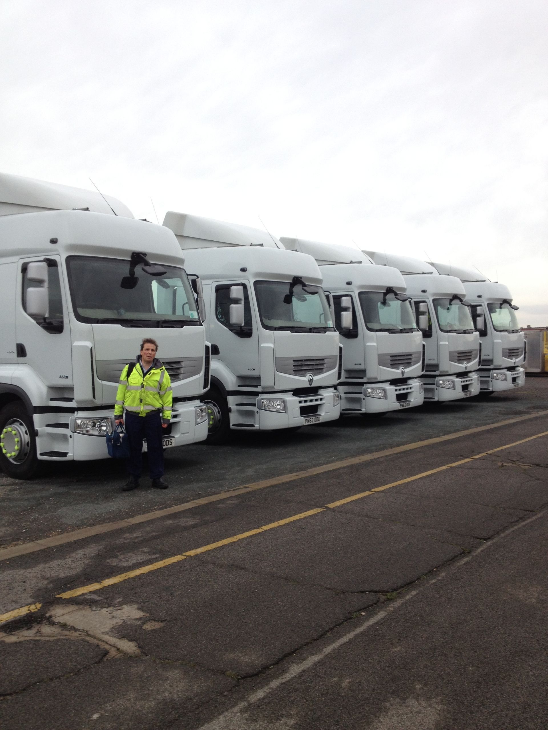 Woman in safety vest standing by a row of white semi-trucks on a paved lot.