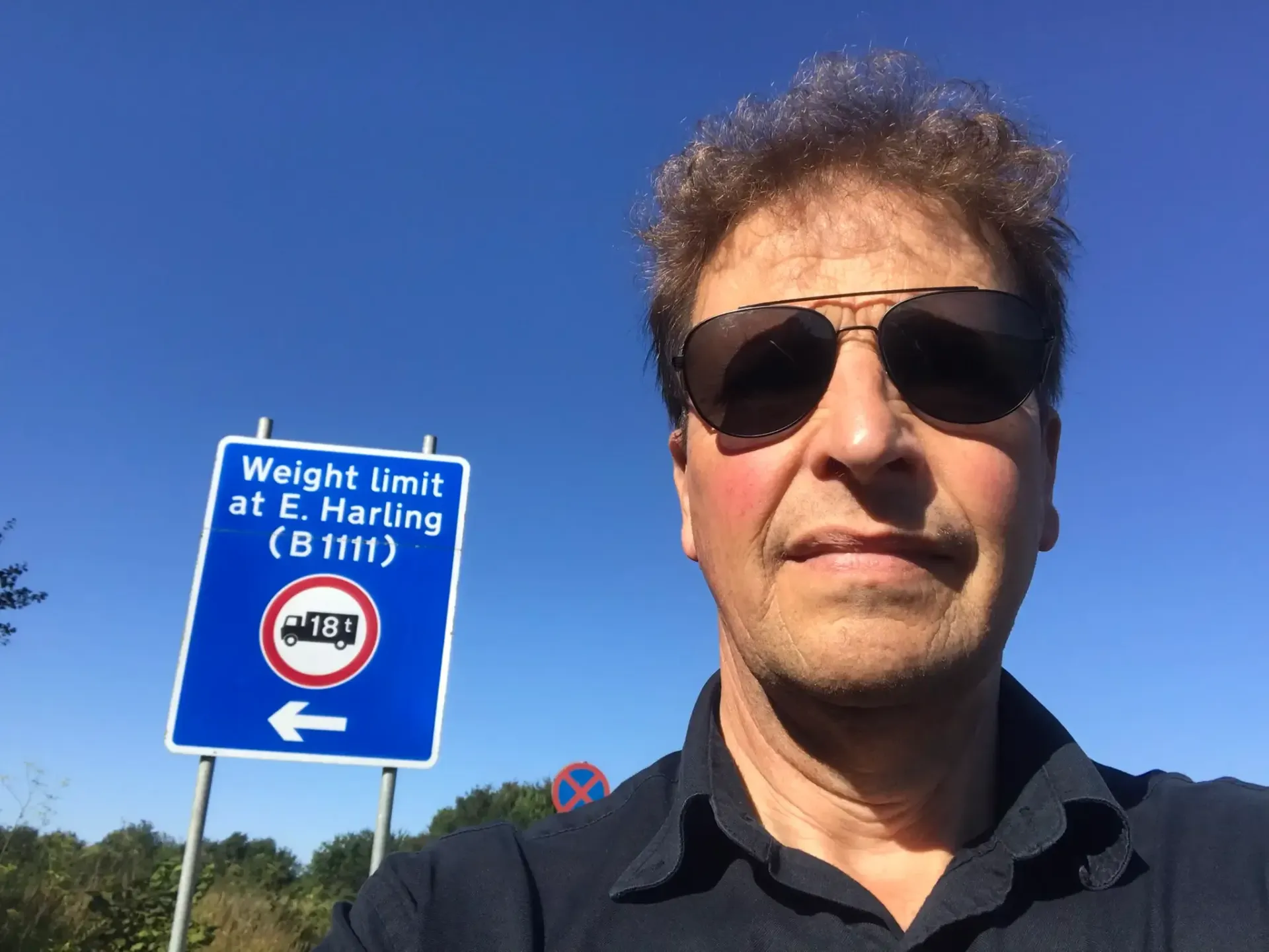 'Harling the Trucker' wearing sunglasses next to a blue weight limit sign with a truck symbol, clear sky in background.