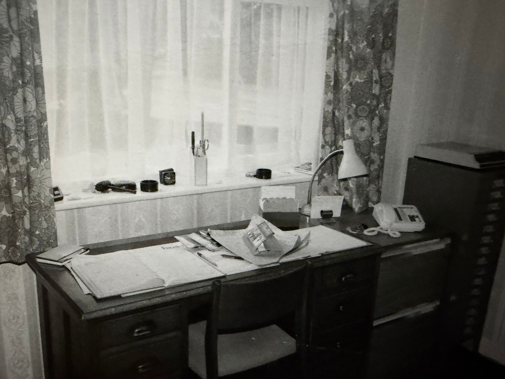 A desk with papers, lamp, phone, and cameras near a window with floral curtains.