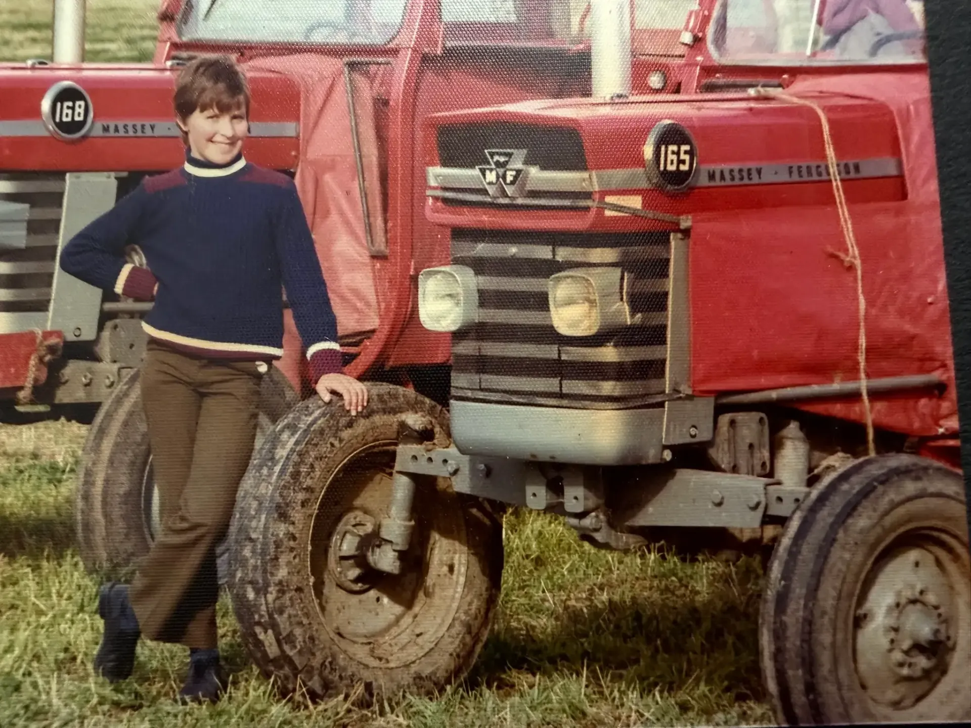 Young person in a sweater leans against a red Massey Ferguson 165 tractor in a field. Another tractor visible behind.