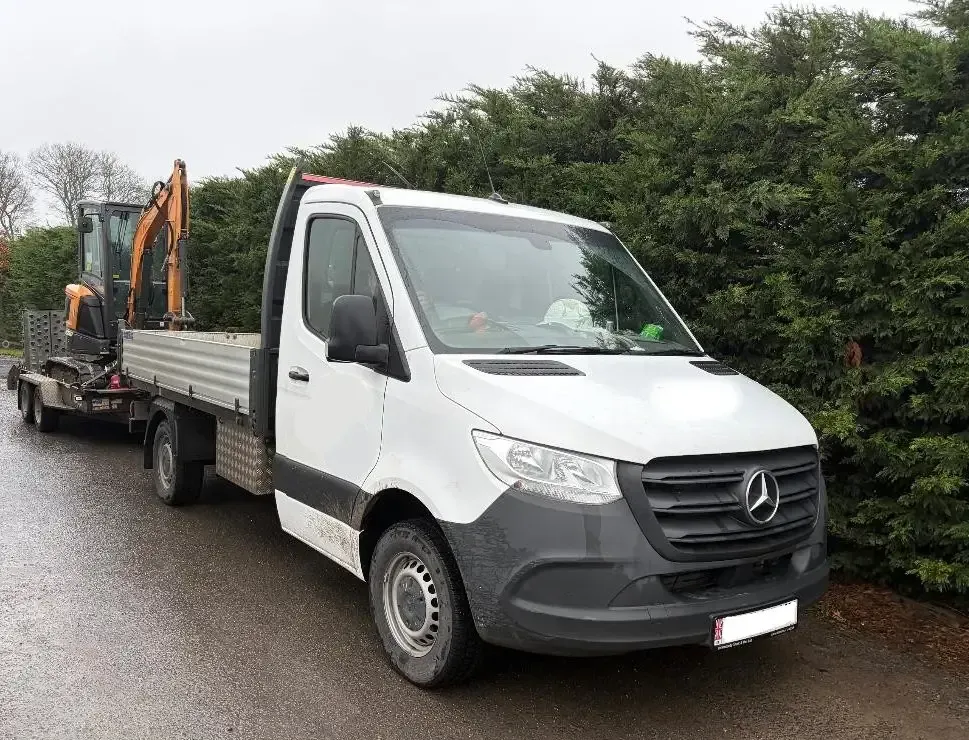 White Mercedes truck with a flatbed pulling a trailer carrying a small excavator on a road next to greenery.