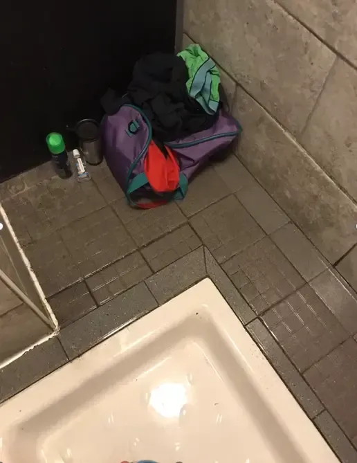 A small purple gym bag with clothing in the corner of a bathroom with a white sink.