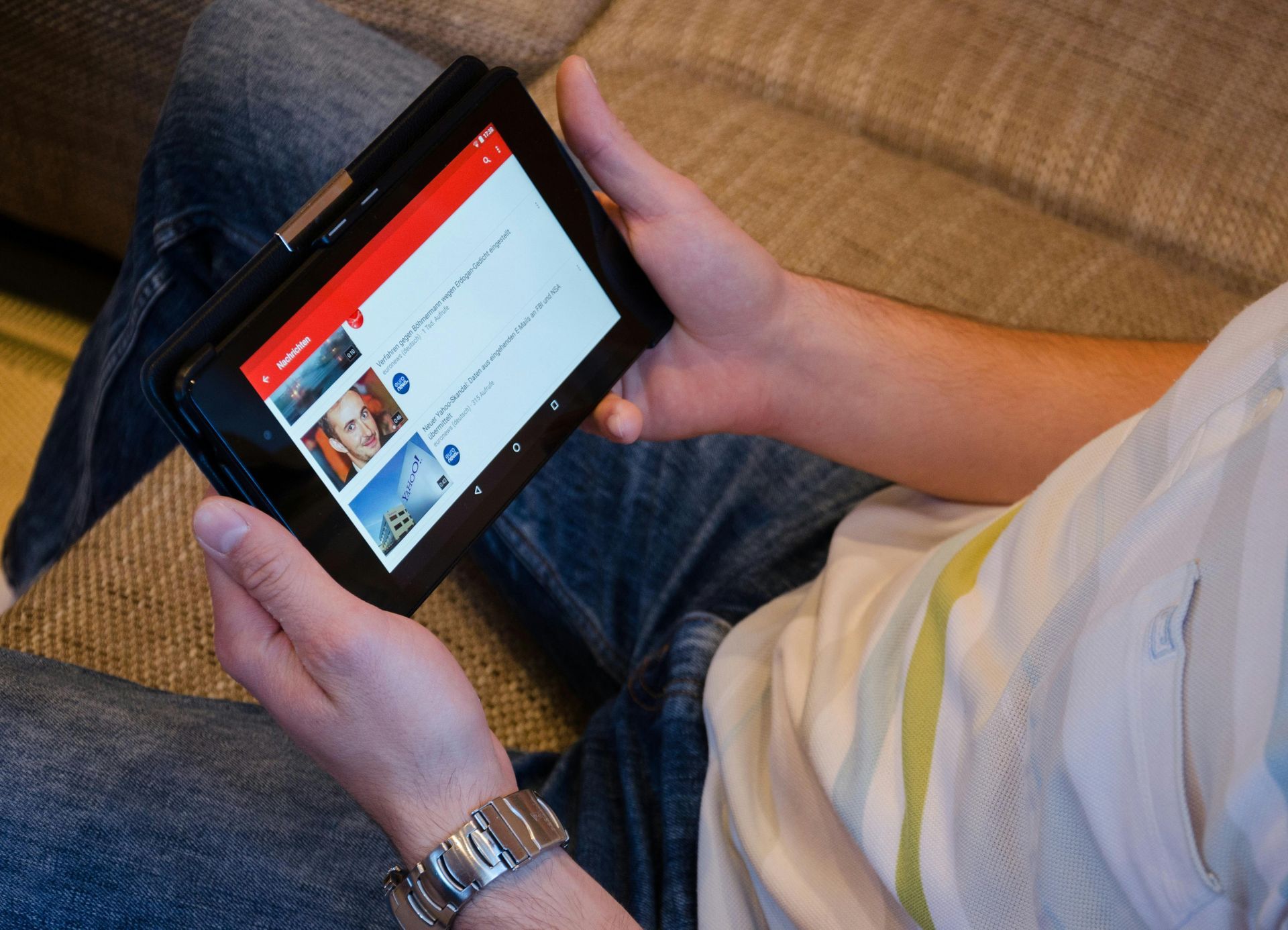 Person sitting, holding a tablet displaying a YouTube list. Red and black interface, watch visible.