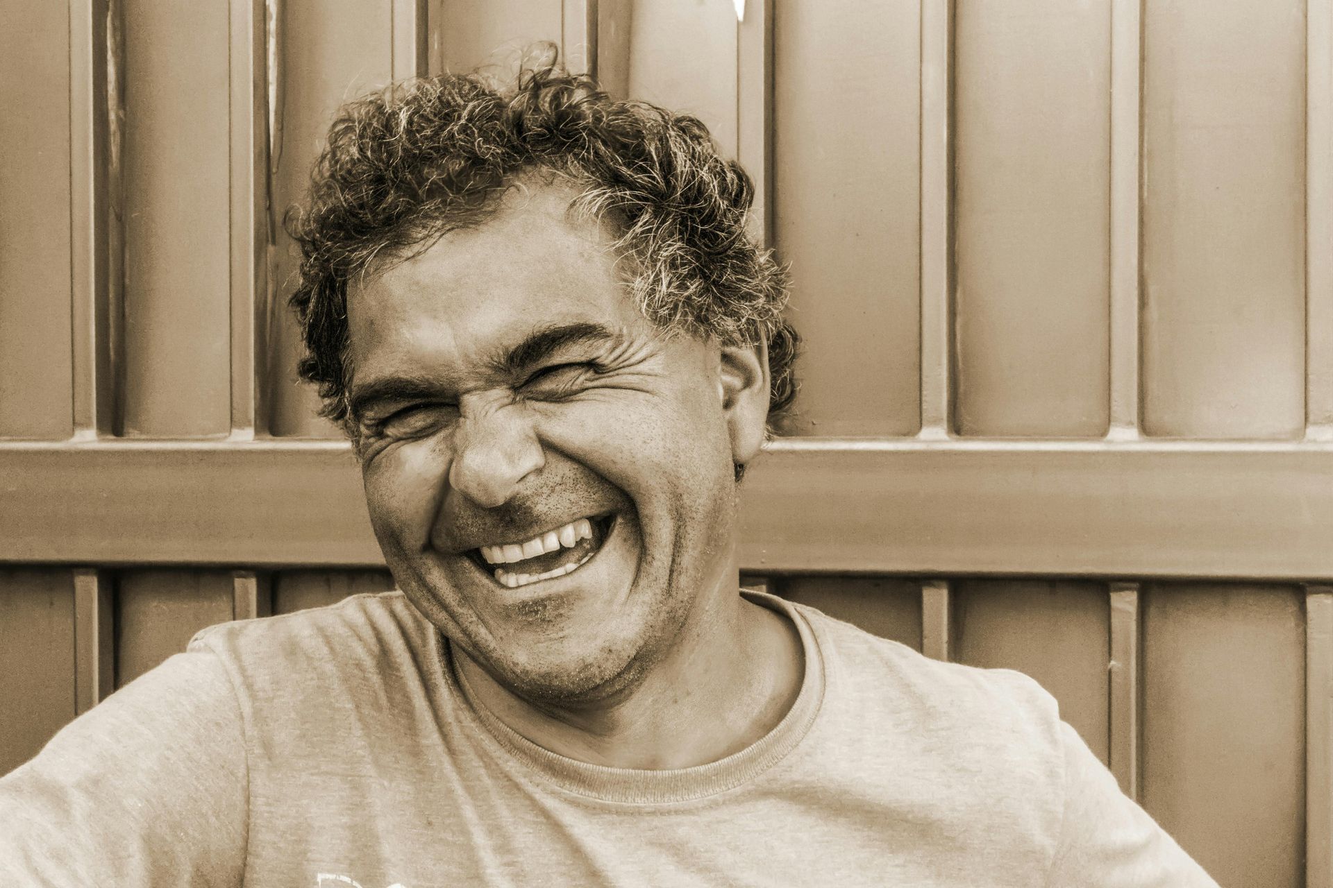Man laughing with eyes closed, in front of a metal gate. Sepia tone.