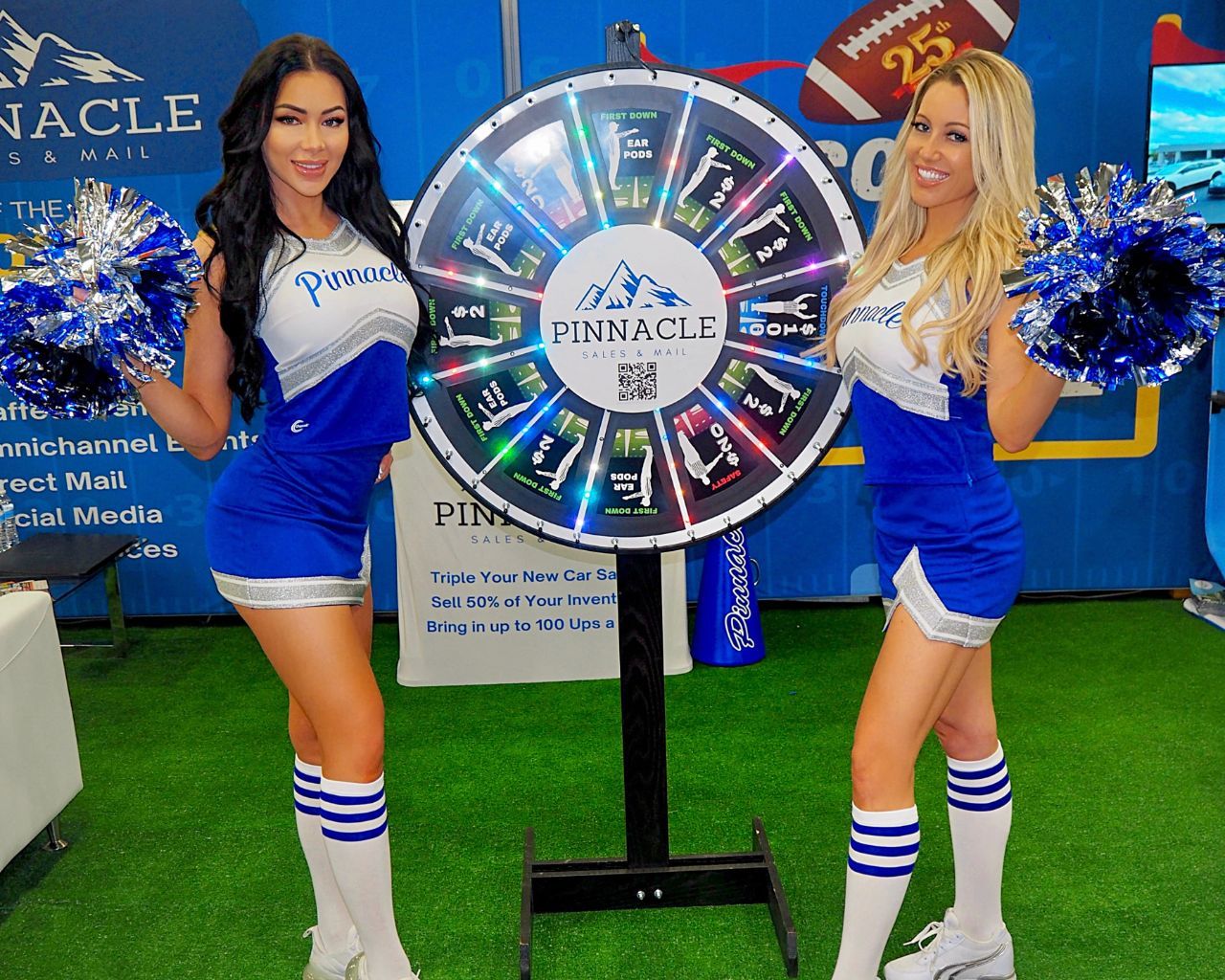 Two models in blue cheerleading outfits with pom-poms stand beside a prize wheel at an event.
