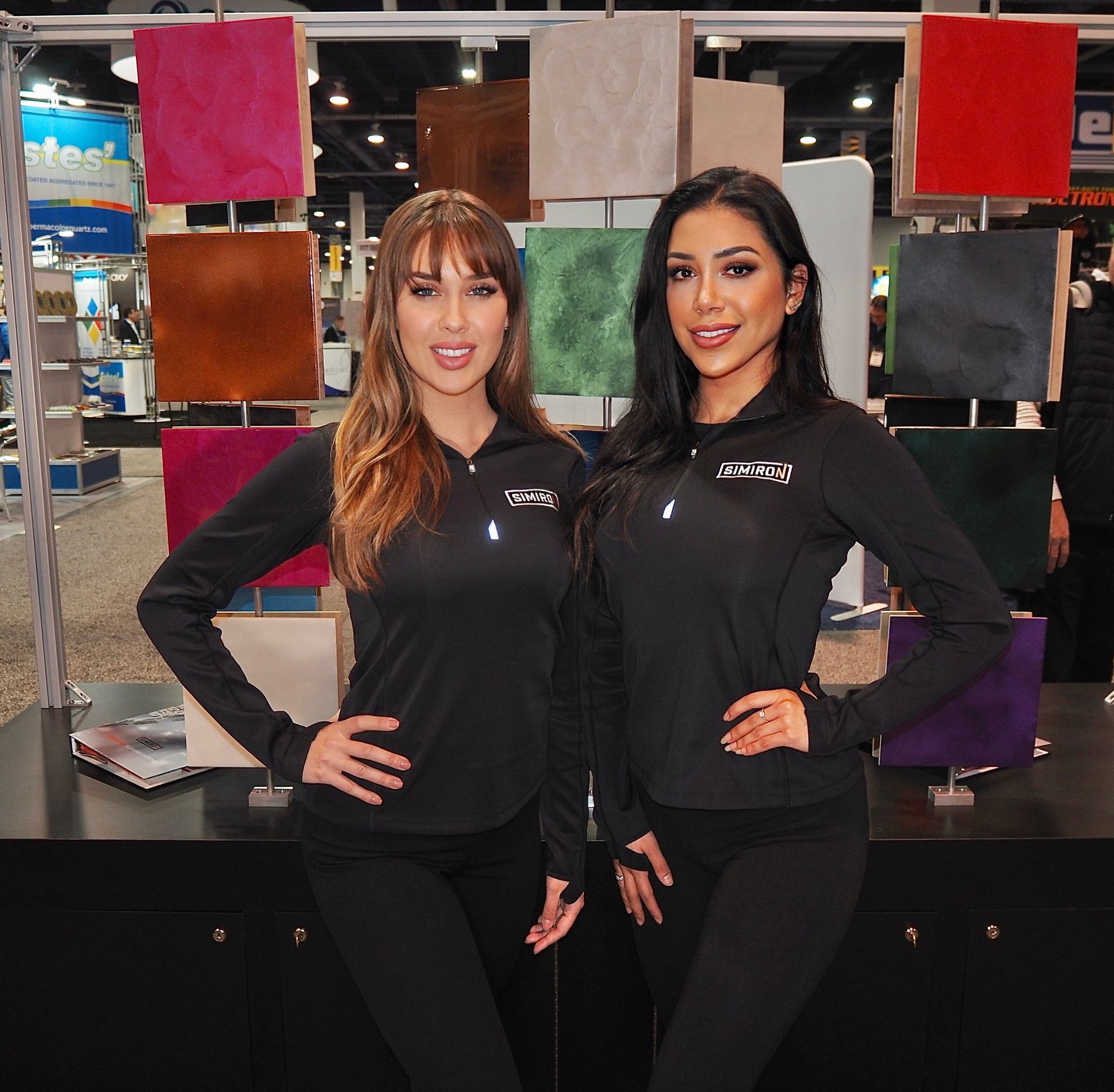 Two trade show models in black shirts and pants pose in front of fabric swatches at a trade show.