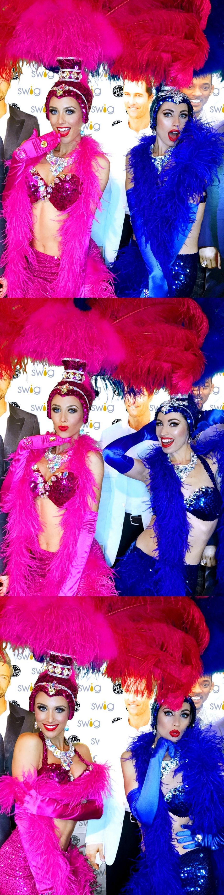 Two showgirls in flamboyant costumes. One in pink, the other in blue, both with feather boas and elaborate headwear.