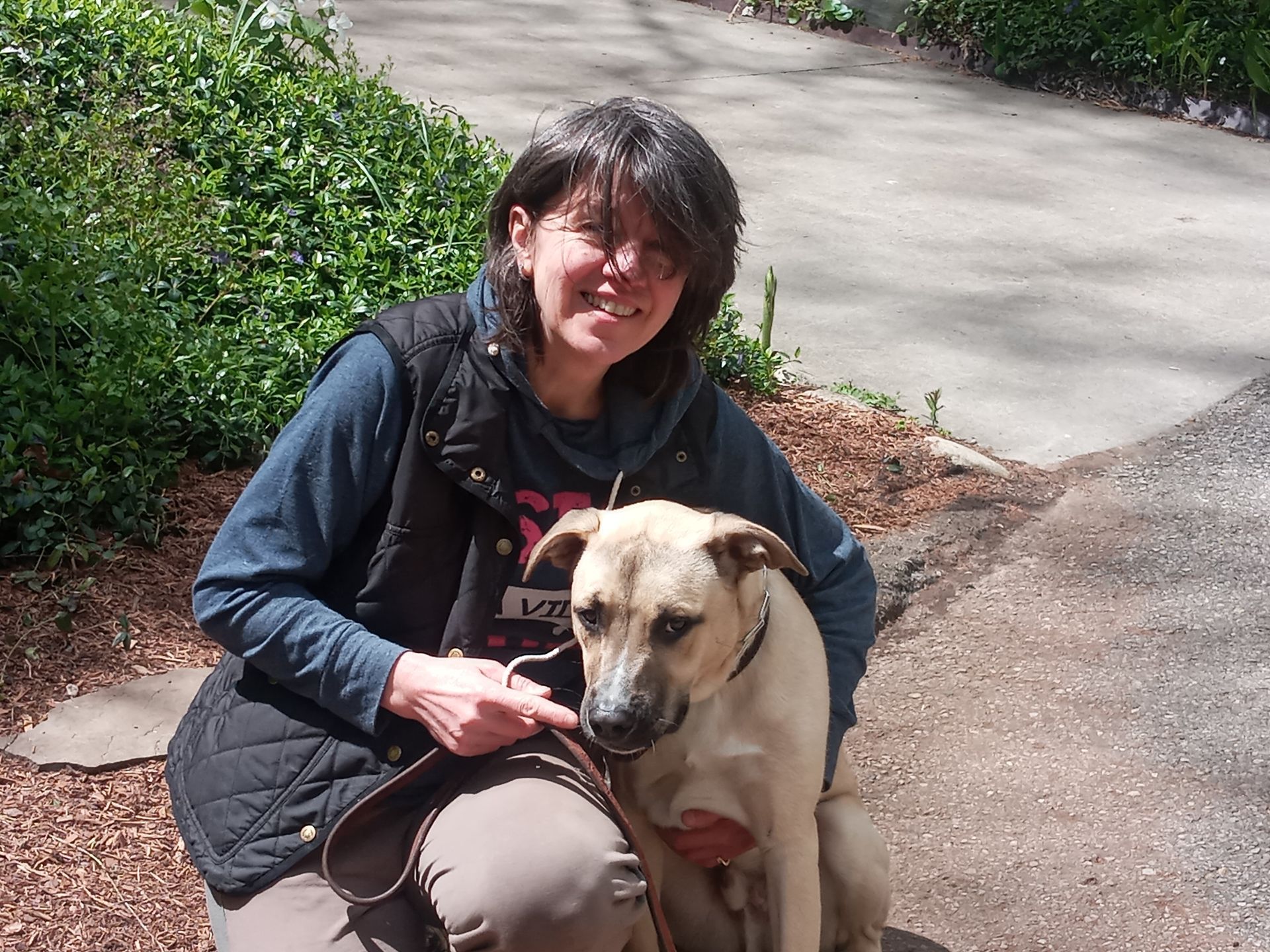 Woman kneels with a tan dog on a path in front of greenery. Both are looking at the camera and smiling.