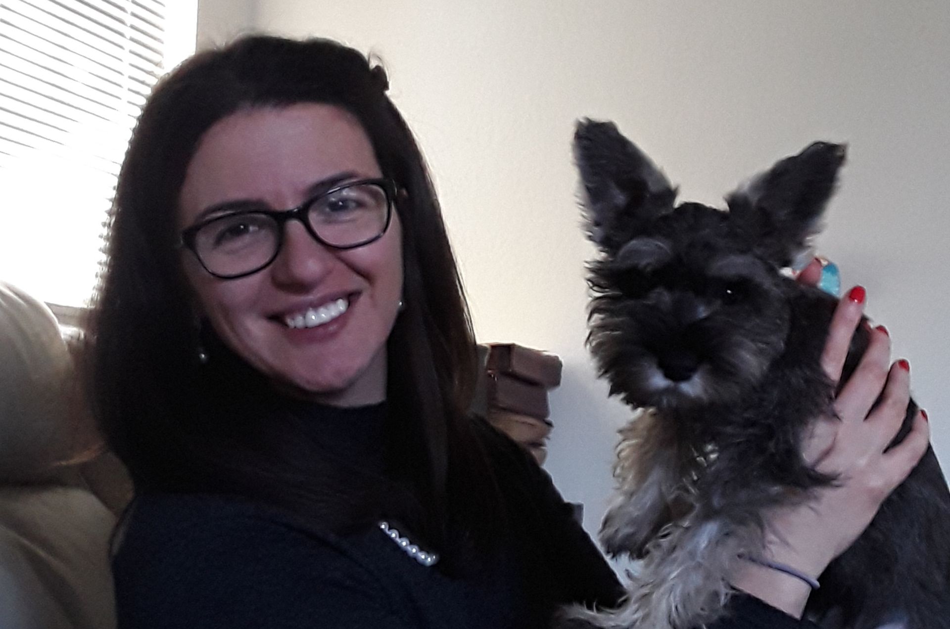 A person with dark hair and glasses smiles while holding a small, dark-haired dog indoors.
