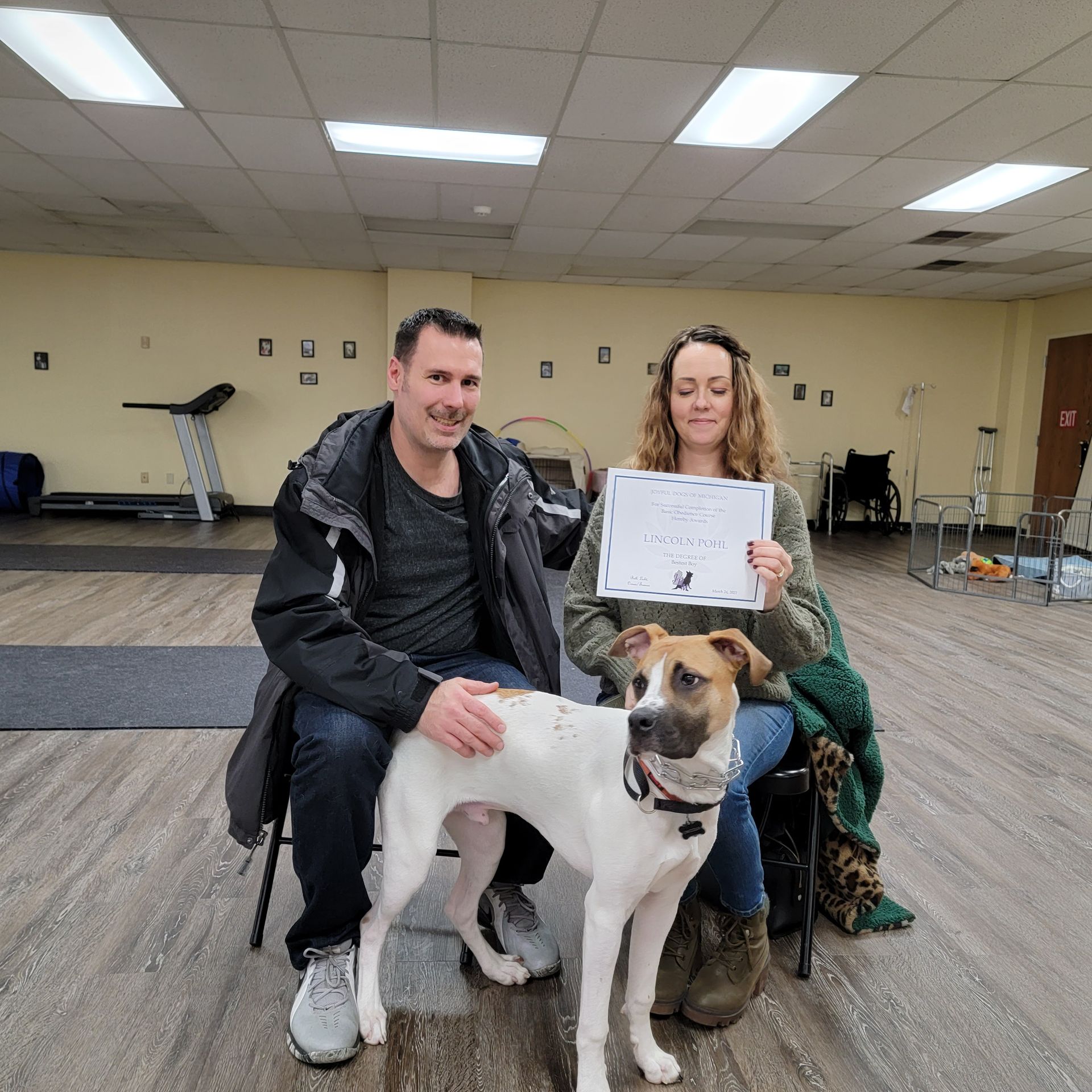 Couple sitting with dog, holding certificate. Indoor setting: exercise equipment visible.