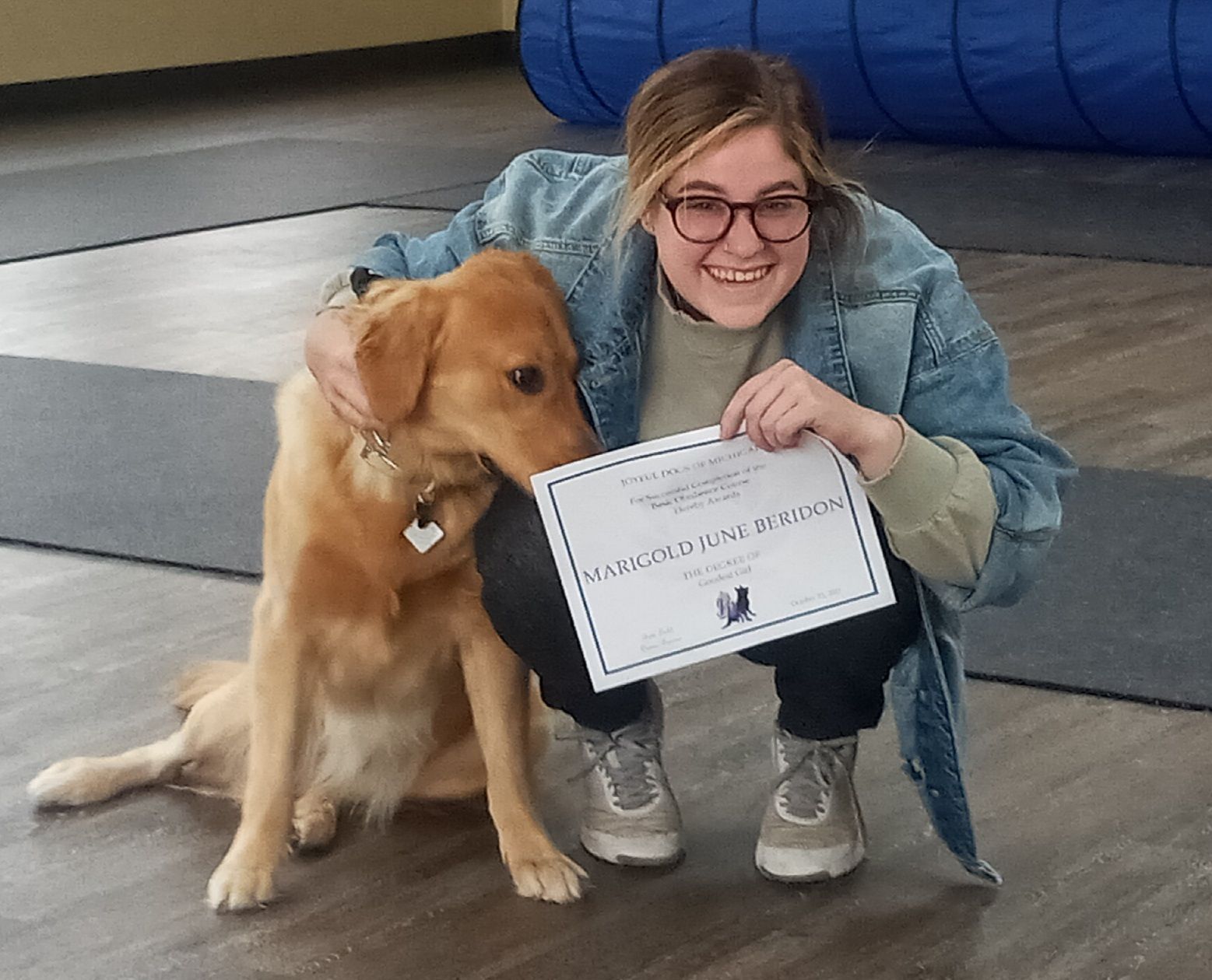 Woman and golden retriever dog holding a certificate. Both smiling, indoor setting, blue tunnel in background.