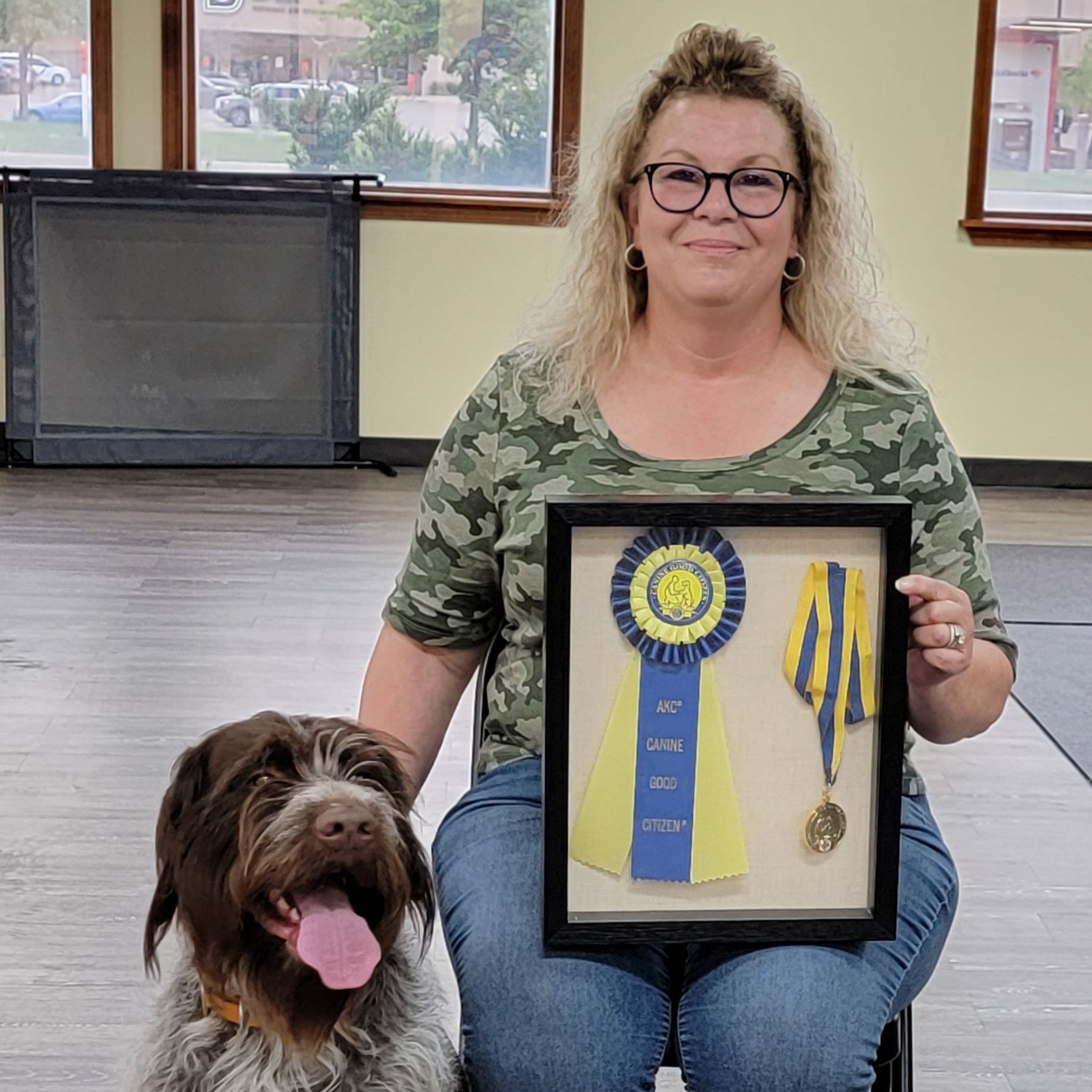 Woman and dog with framed ribbon and medal. Woman smiles, wearing glasses and camo shirt. Dog sits beside her.