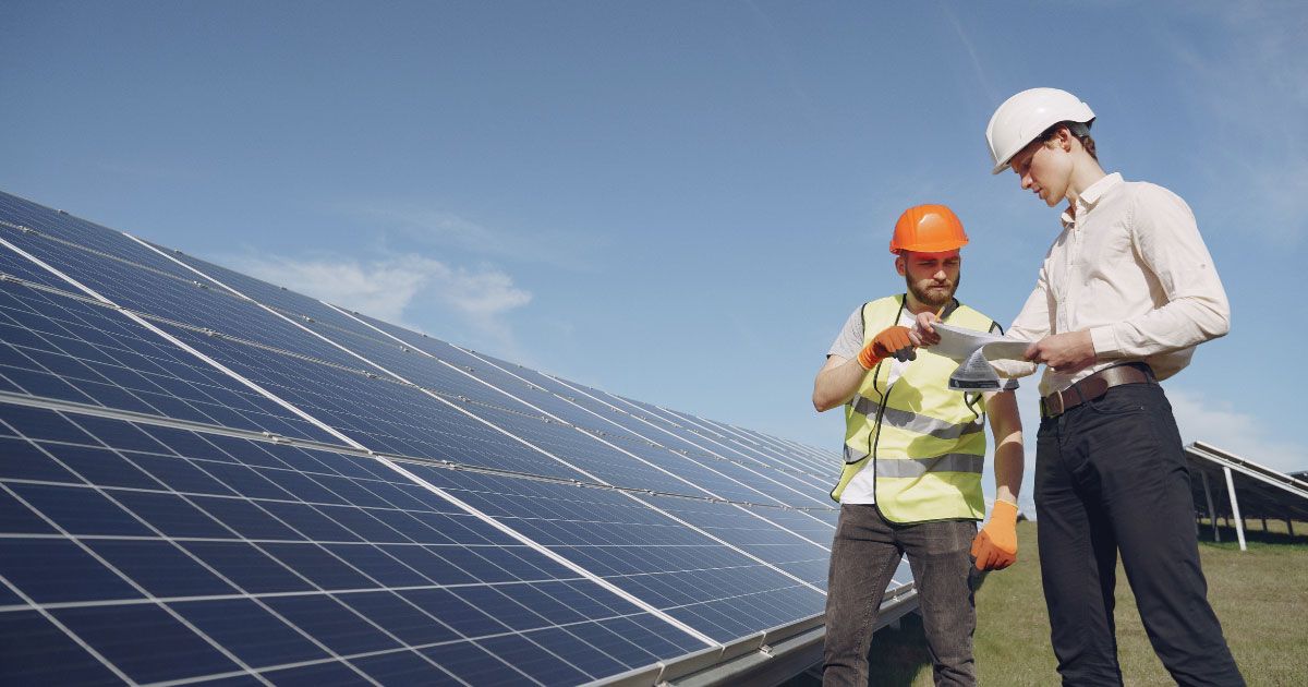 Two men are standing in front of a field of solar panels looking at a tablet.