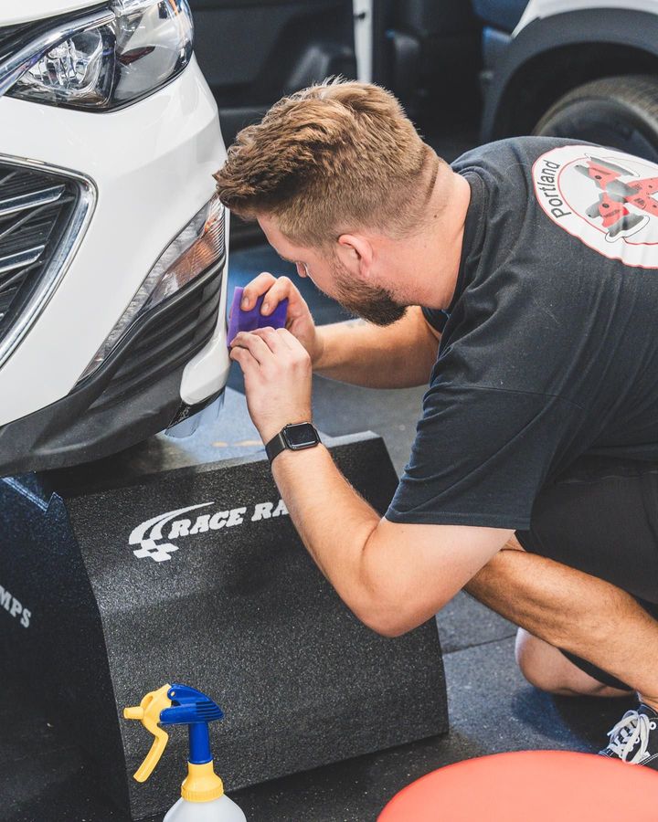 Person polishing the front bumper of a white car with a cloth in a garage or auto shop