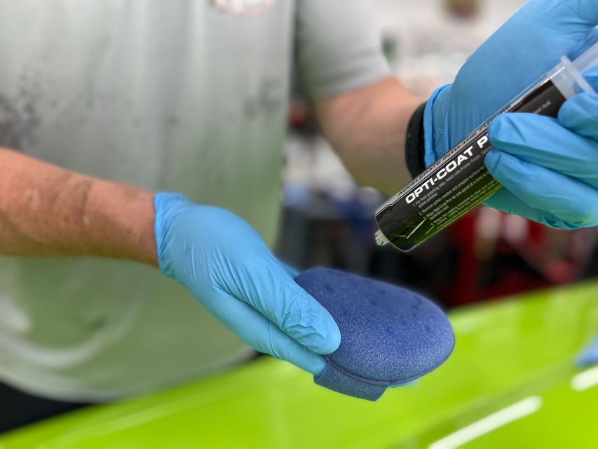 Gloved hands hold a blue sponge and a bottle of cleaning solution over a green surface.