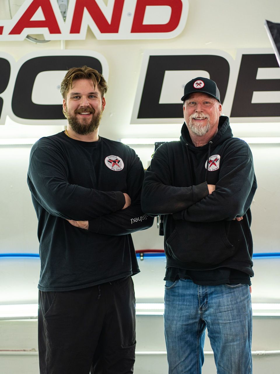 Two men in black shirts and jeans standing with arms crossed in front of a large sign.