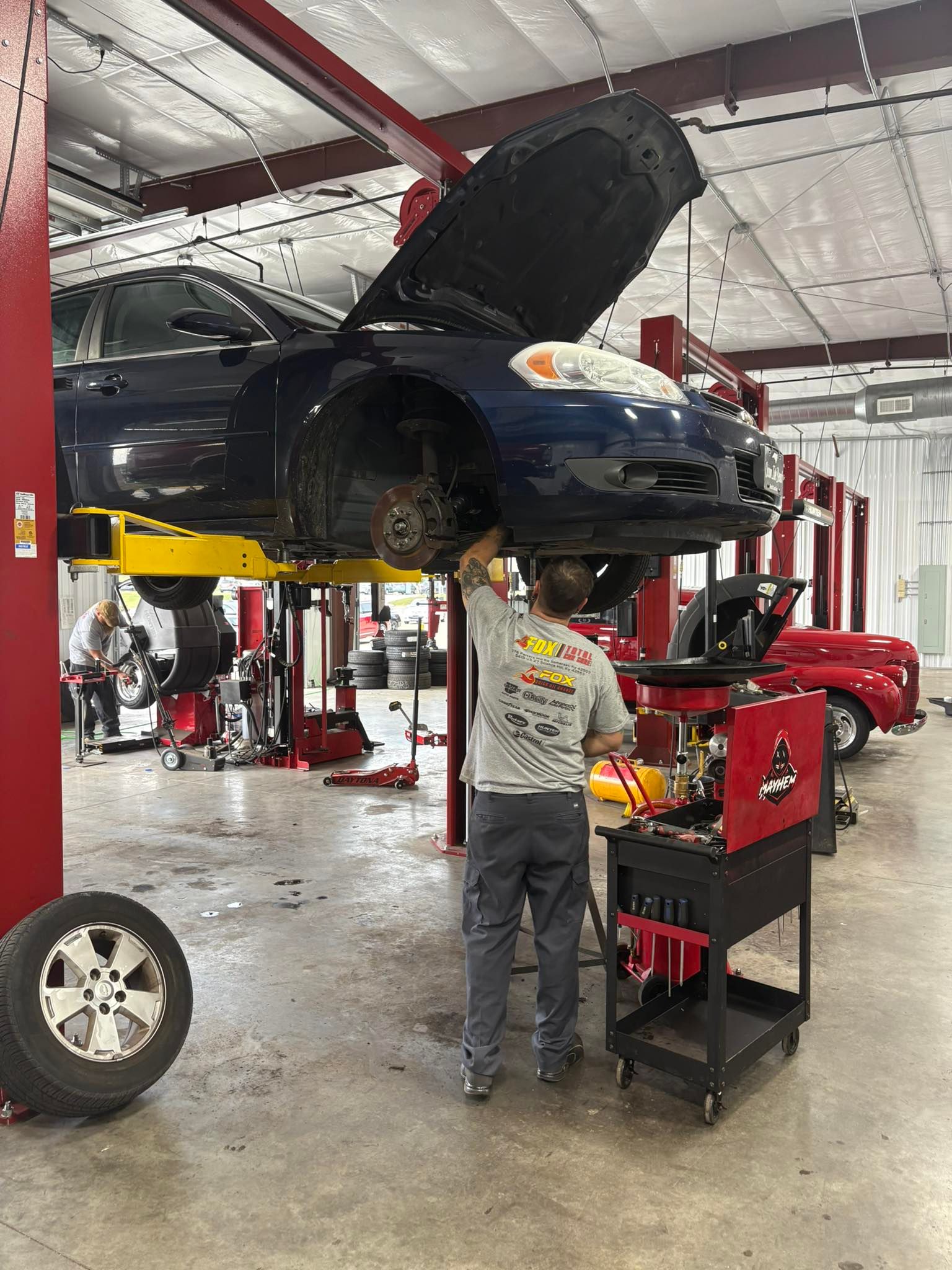 Mechanic working on a blue car raised on a lift in a garage. Tools on a cart nearby.