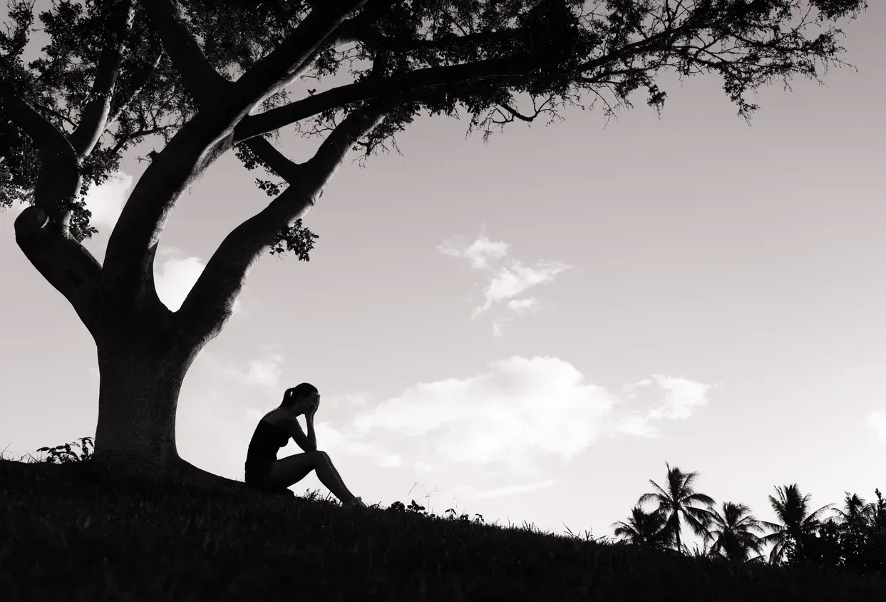 Woman sits under a tree, head in hands, looking down. Black and white, on a grassy hill.