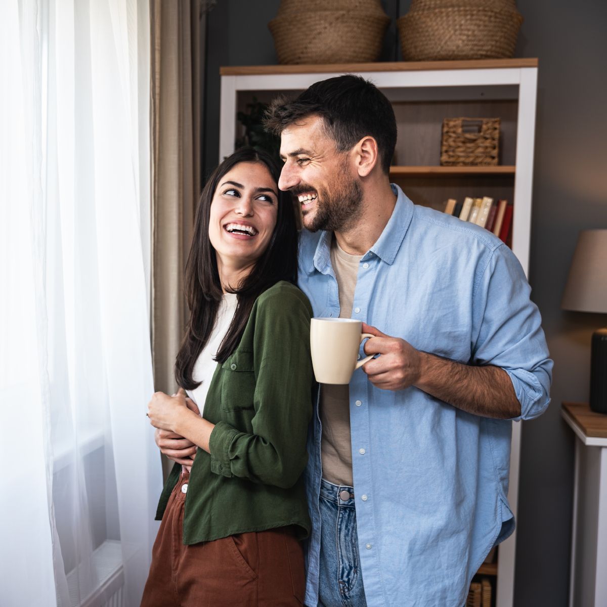 A man and a woman are standing next to each other in front of a window holding a cup of coffee.