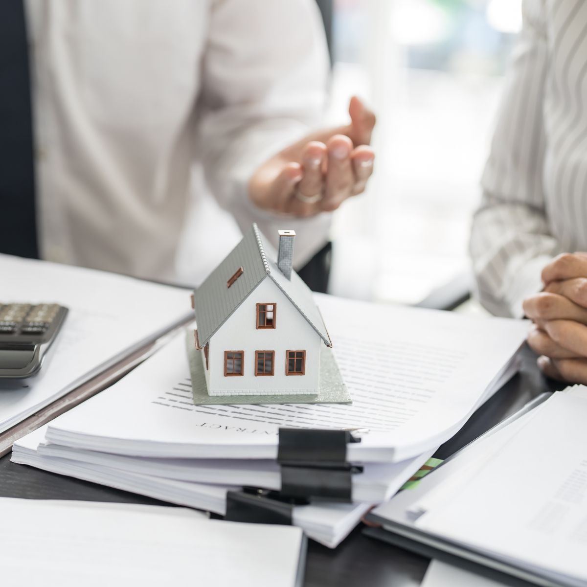 Two people are sitting at a table with a model house on top of a pile of papers.