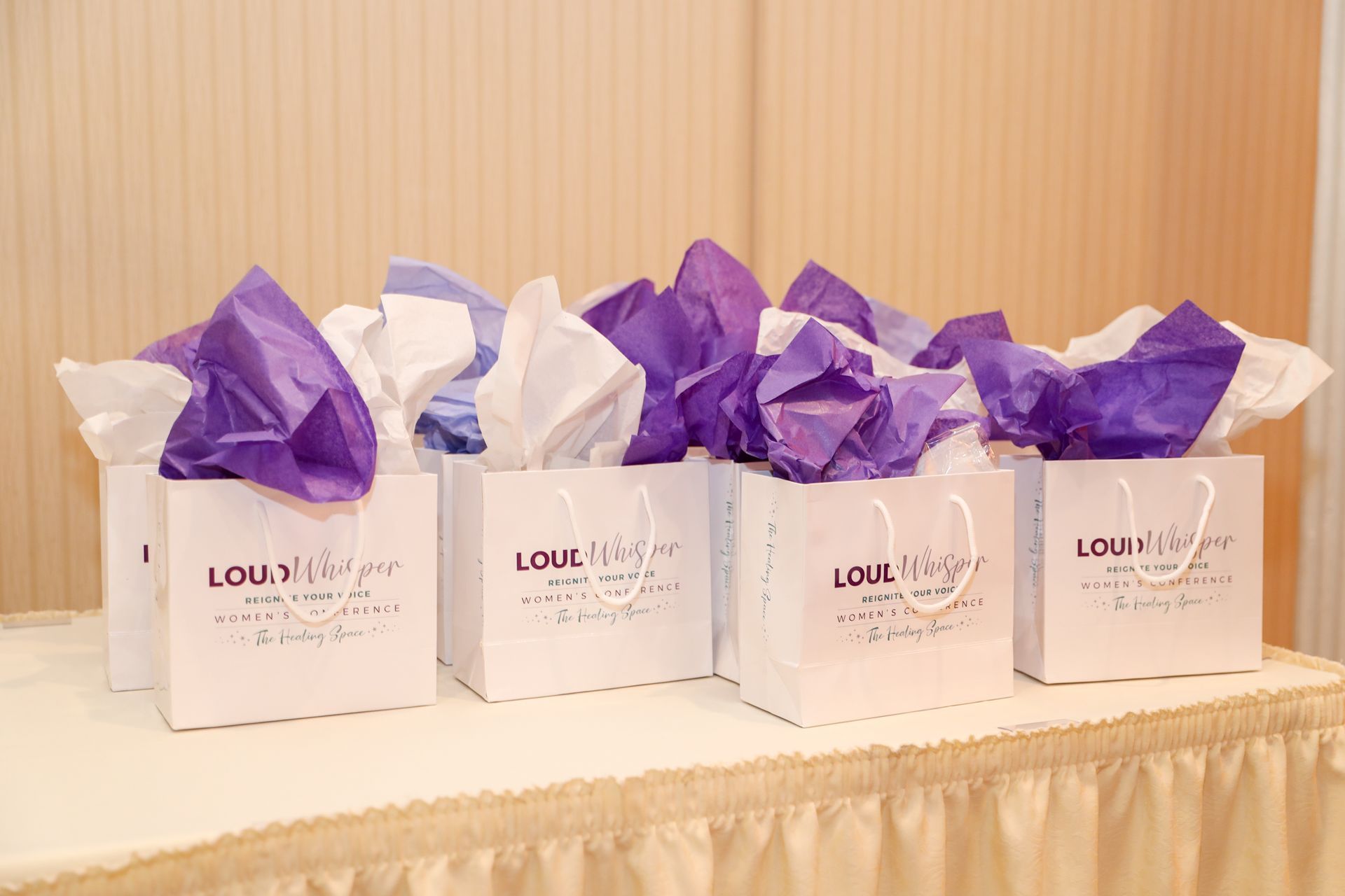 Four white gift bags with purple and white tissue paper on a table.