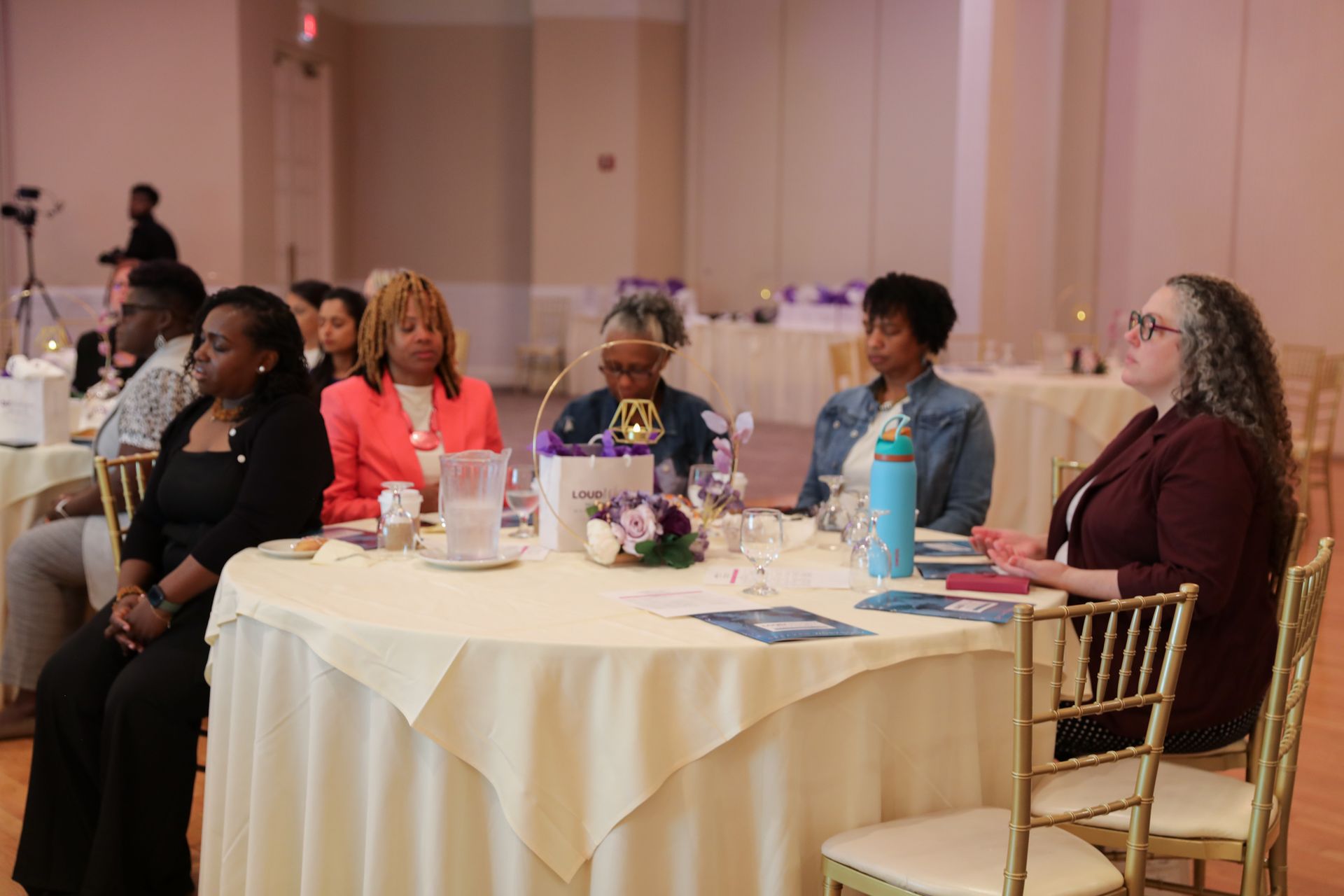 People seated around a table at an event. Attendees listen, some look at the speakers. Bright room.