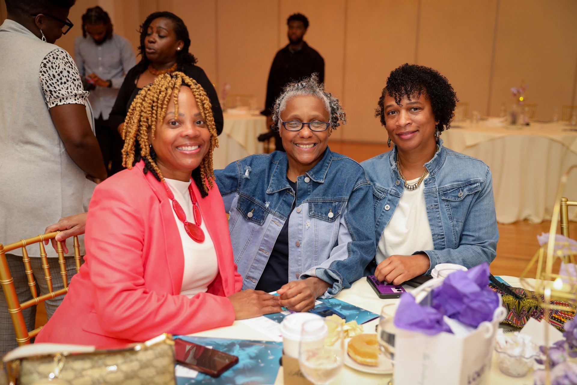 Three women smiling at a table, one in red blazer, others in denim jackets, at an event.