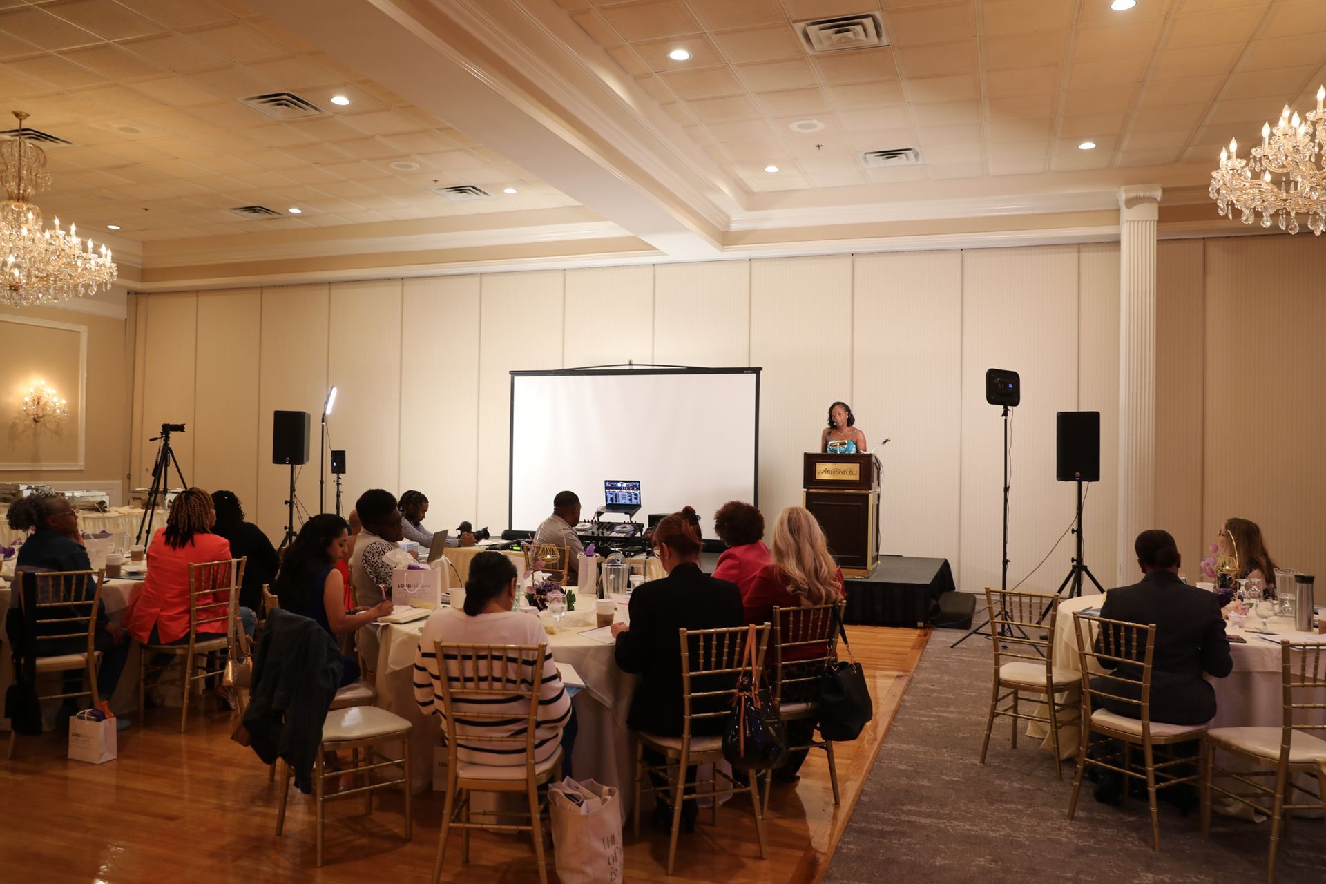 People at tables listen to a speaker at a conference. A screen is behind the speaker.