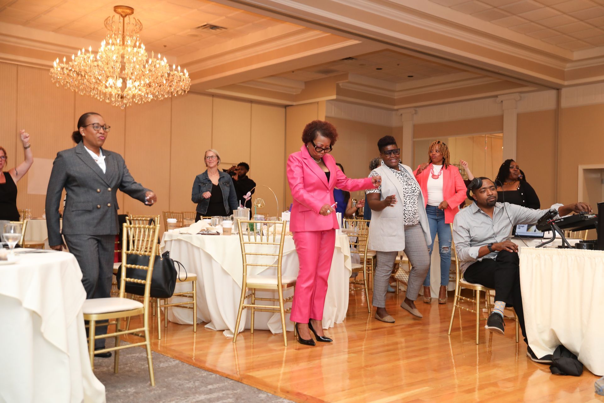 People dancing in a ballroom; a woman in a pink suit leads the group.