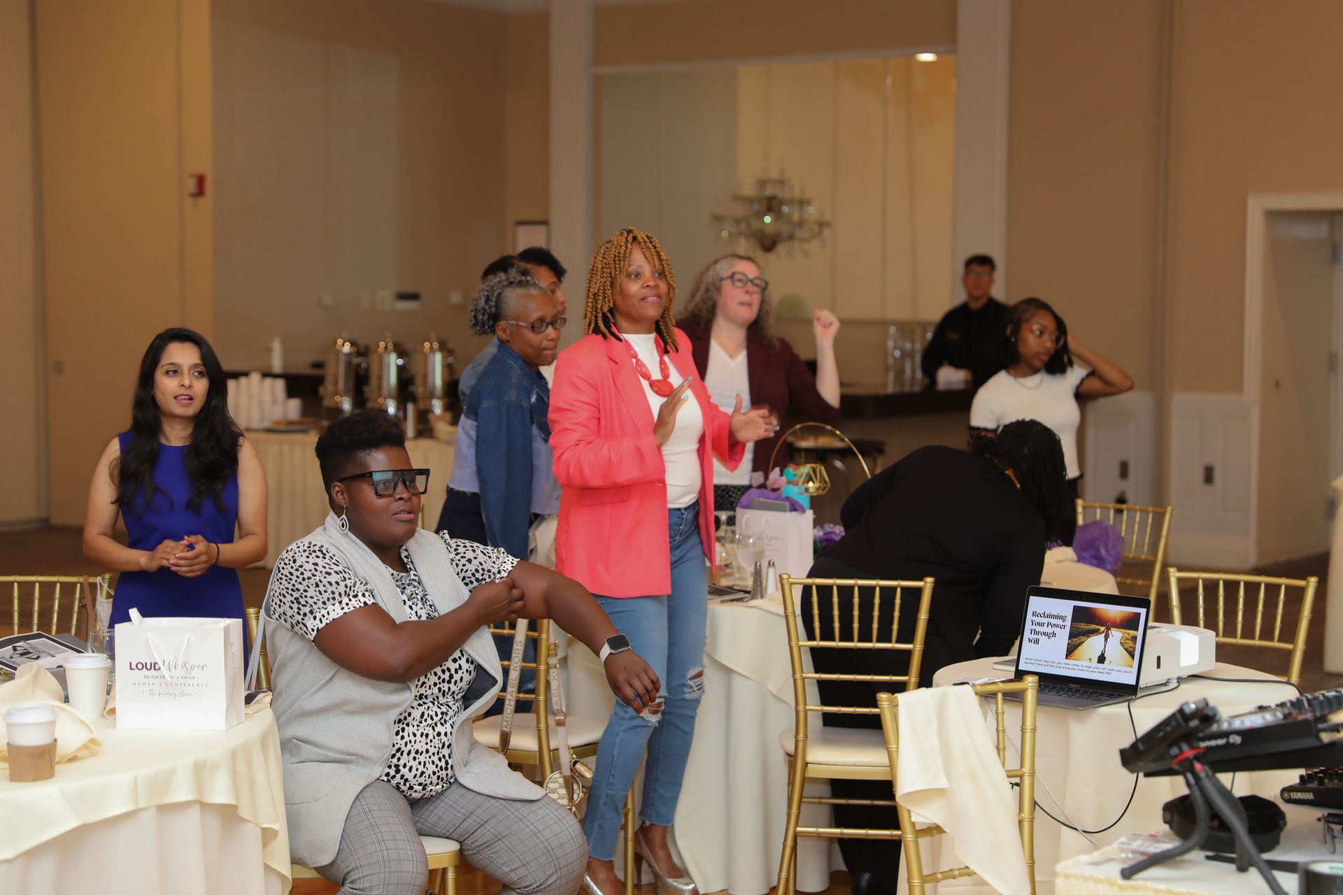 People dancing and interacting at an event, with tables, gold chairs, and a buffet in a bright room.