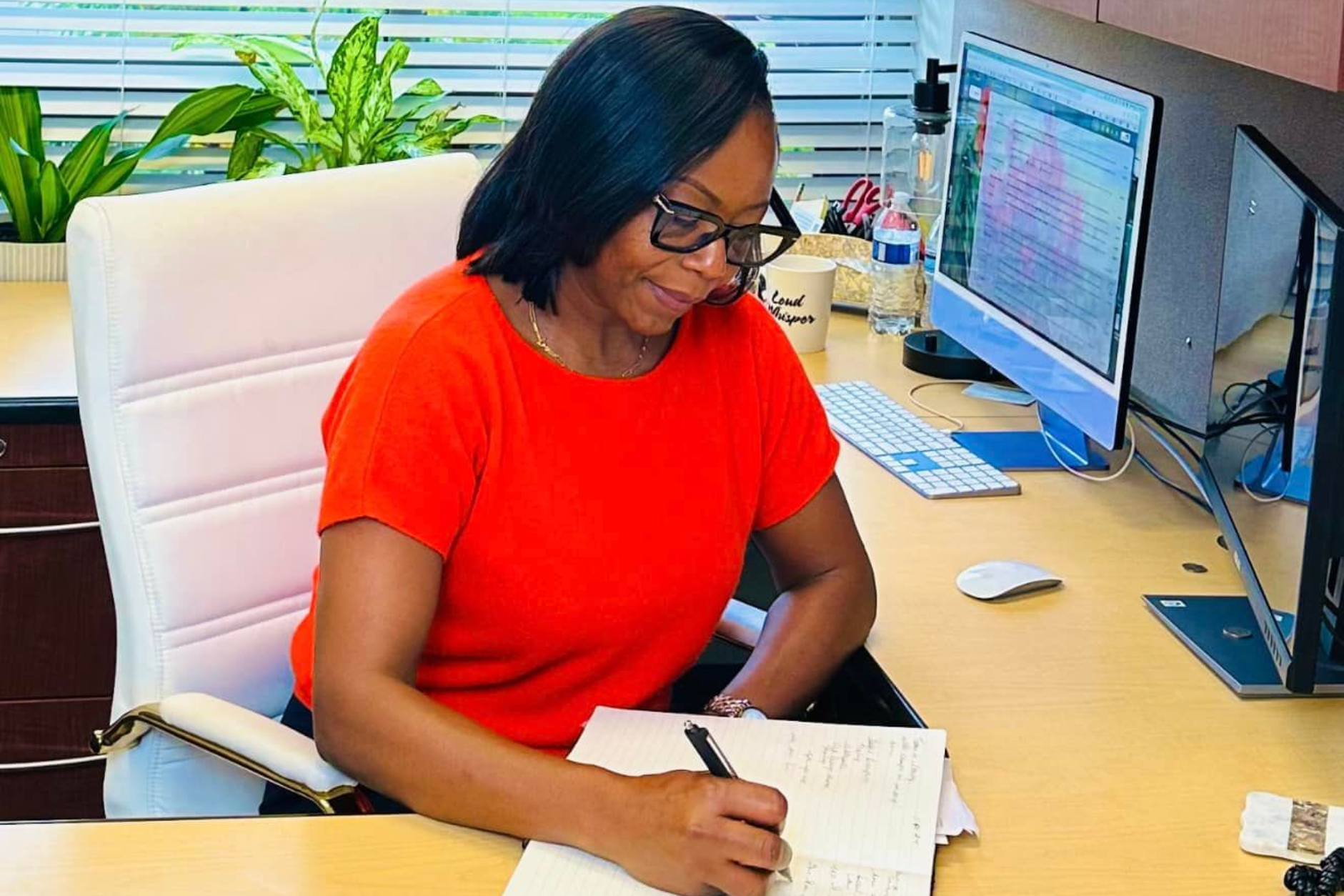 Woman in orange shirt writing at a desk with two computer screens and a plant.