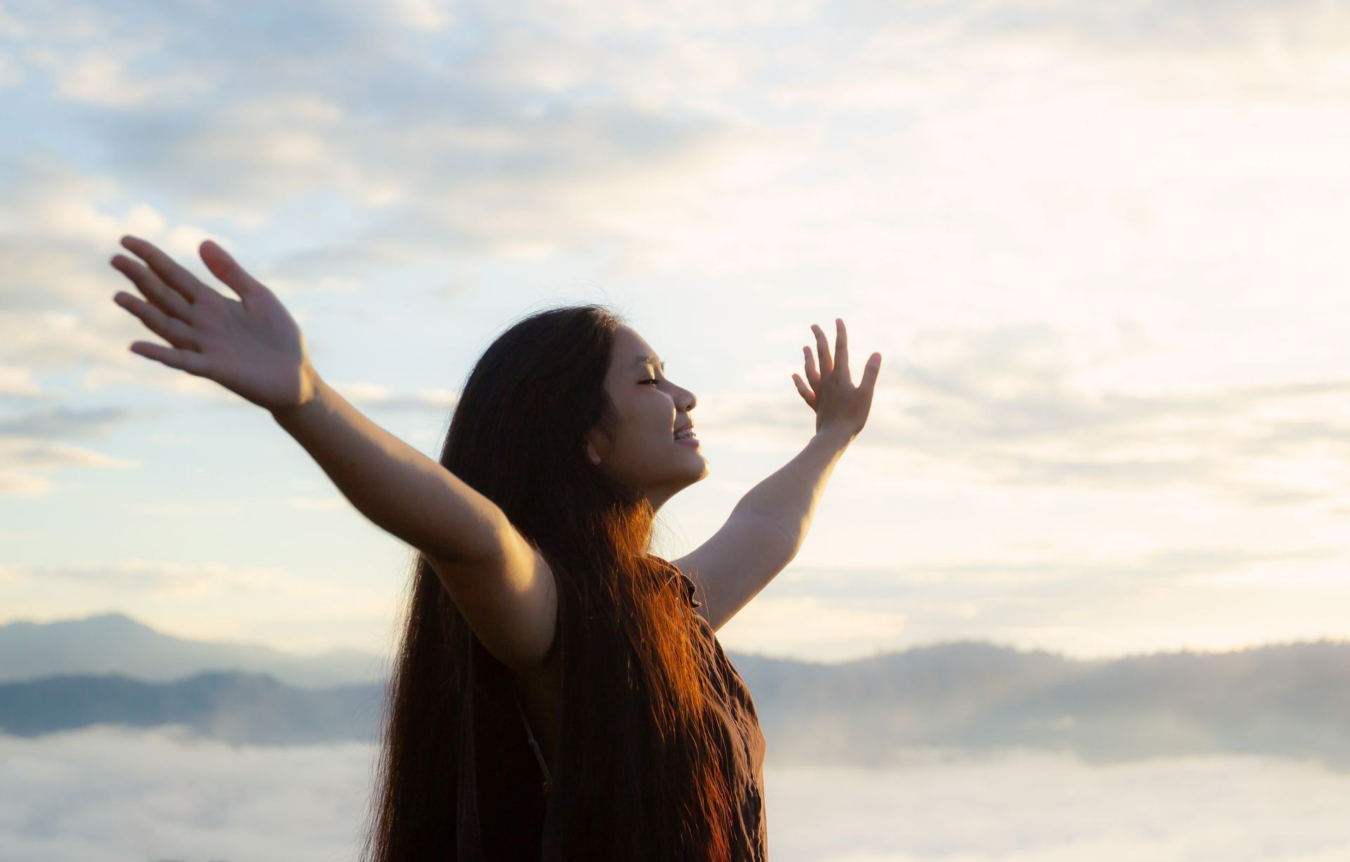 Woman with arms outstretched, smiling, facing the sunlit sky above mountains.