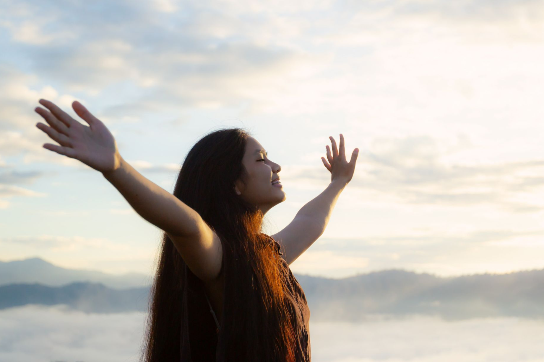 Woman with arms outstretched, smiling, facing the sunlit sky above mountains.
