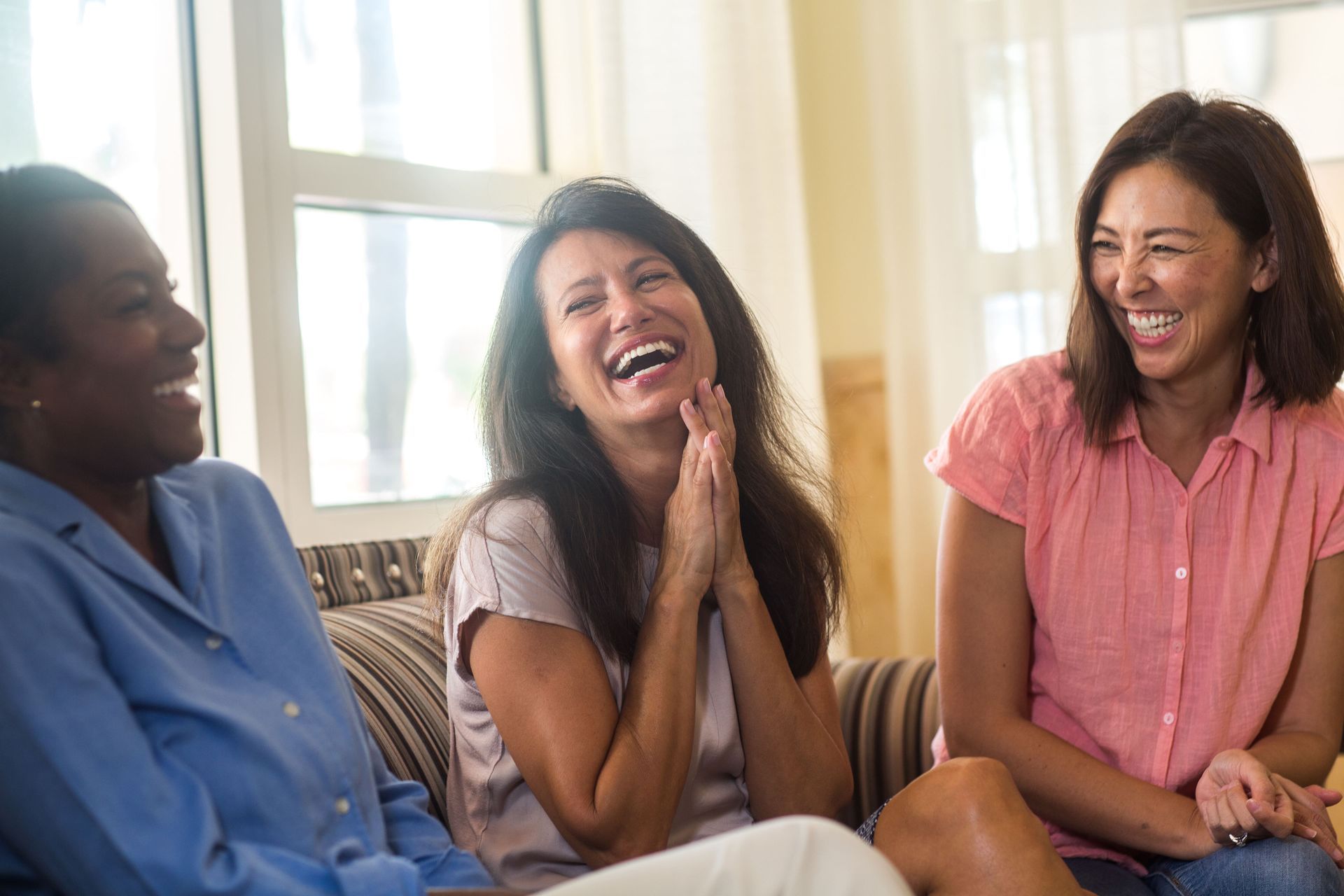 Three women laughing together on a couch near a window.