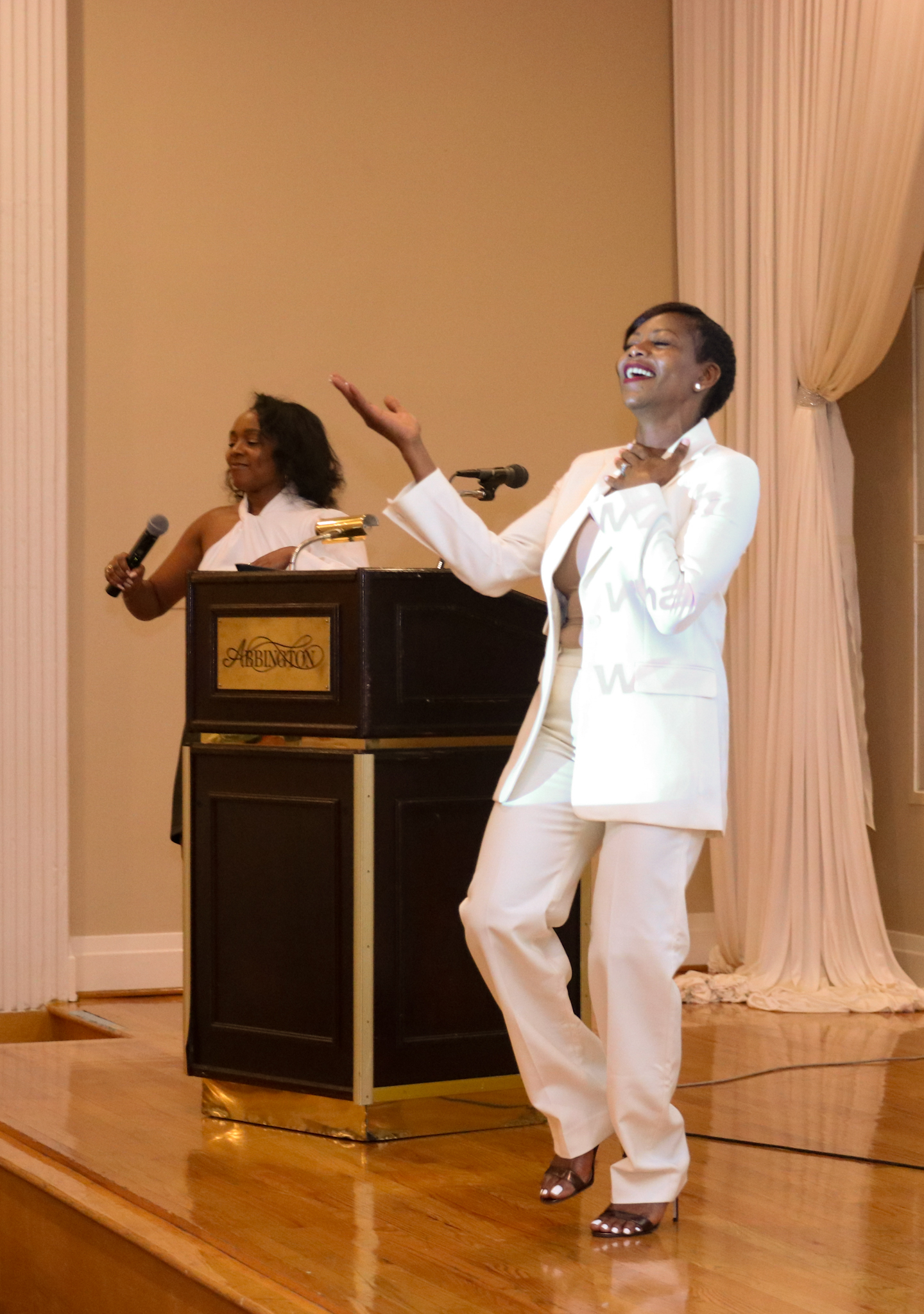Woman in white suit singing, arms raised, on stage; another woman singing behind podium.