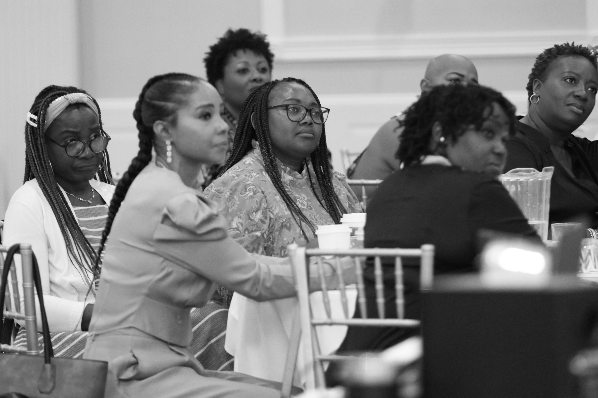 Attendees at a meeting, seated at tables. Some looking forward, some with neutral expressions. Indoors, black and white.