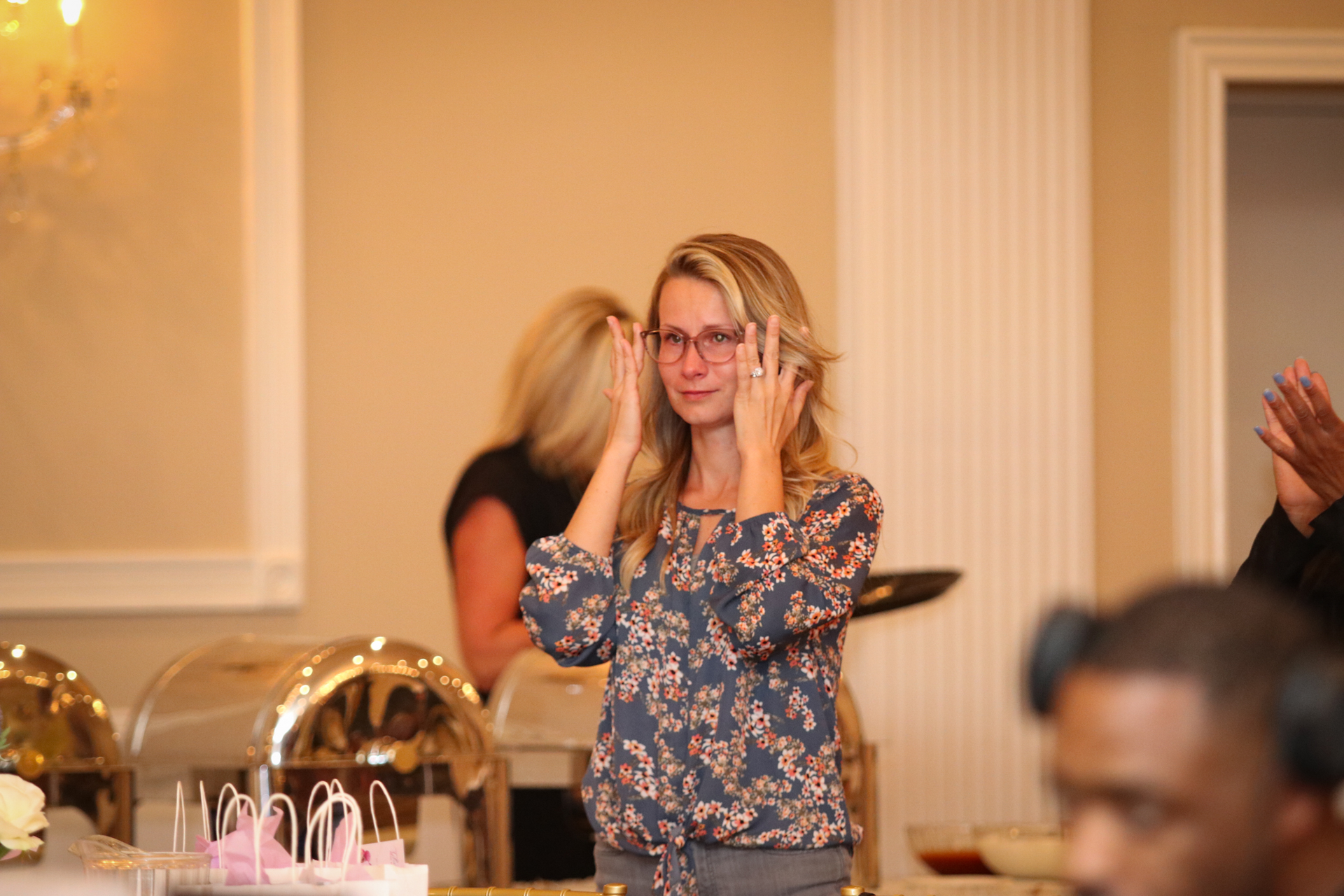 Woman in floral top adjusts glasses, emotional at event. Buffet in background.